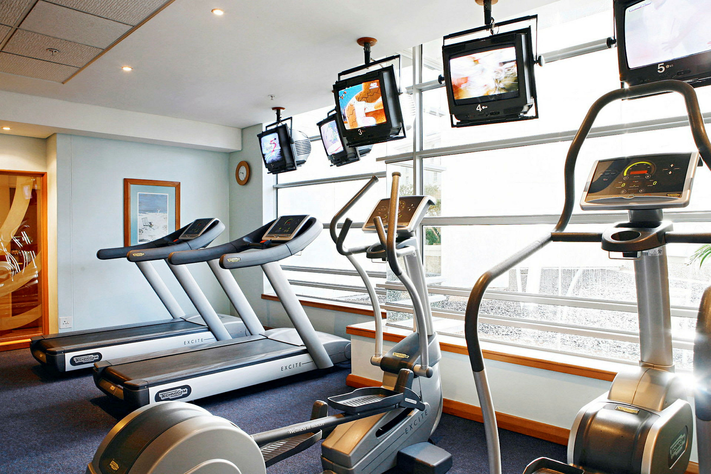 Billede af et fitnesscenter med træningsmaskiner såsom løbebånd og elliptical maskiner samt tv-skærme ved vinduerne. (AI-genereret tekst)