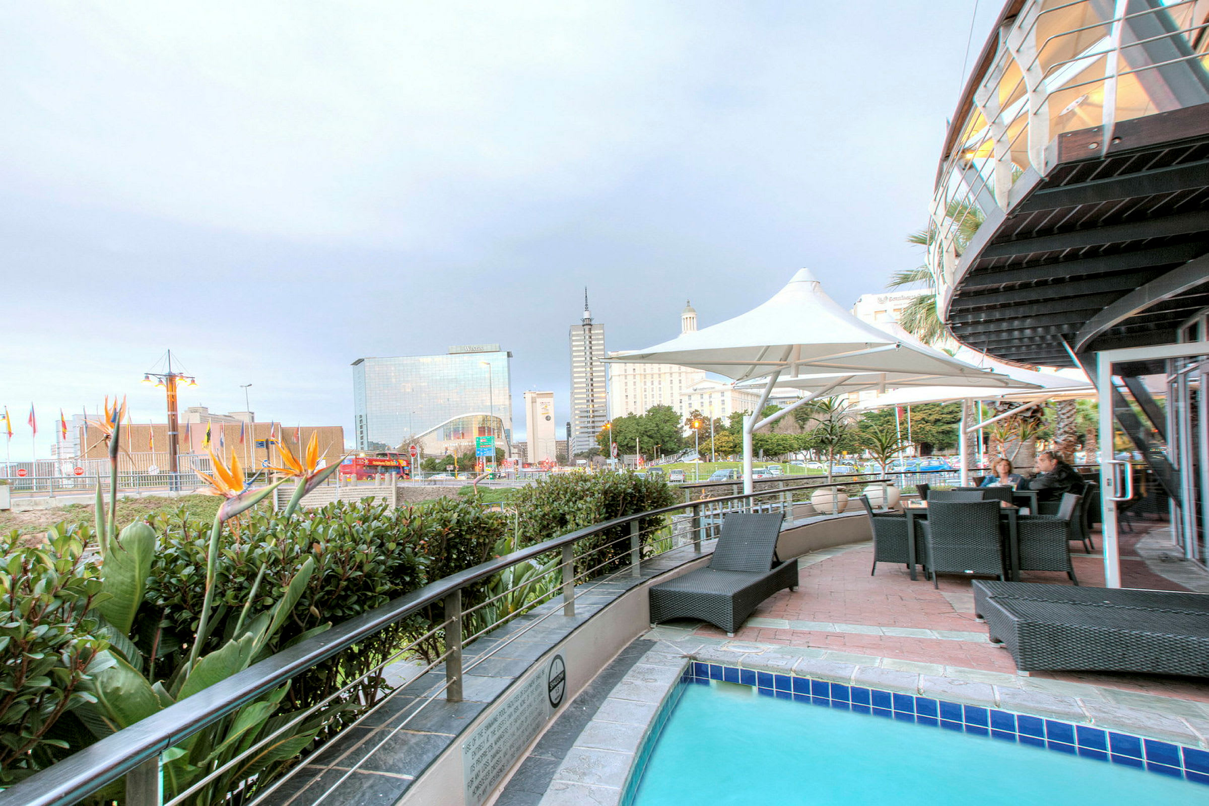En udendørs terrasse med pool, omkringliggende grønne områder og udsigt til moderne bygninger i baggrunden. (AI-genereret tekst)