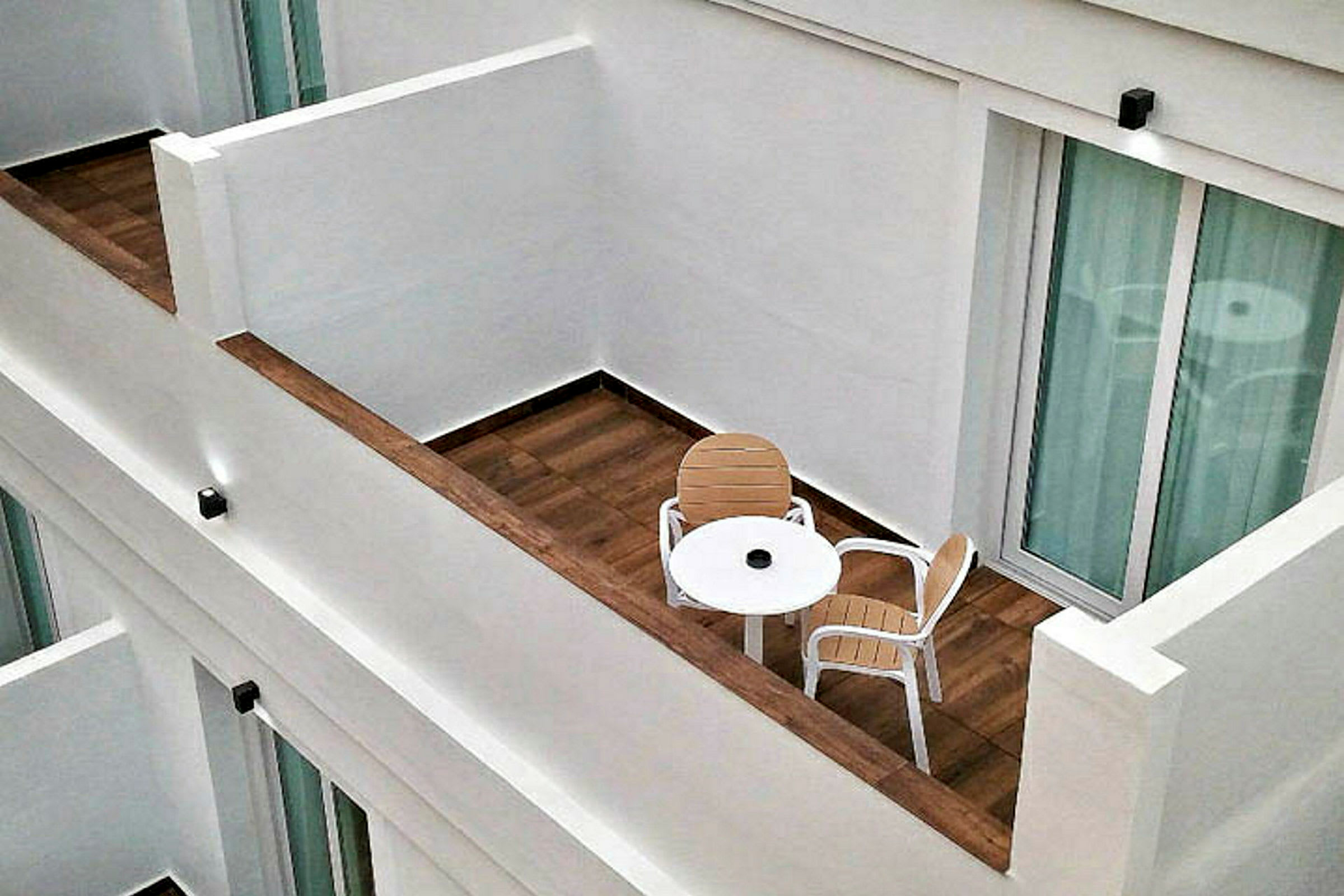 Billedet viser en balkon med trægulv, et rundt hvidt bord og to stole uden for en glas skydedør. (AI-genereret tekst)