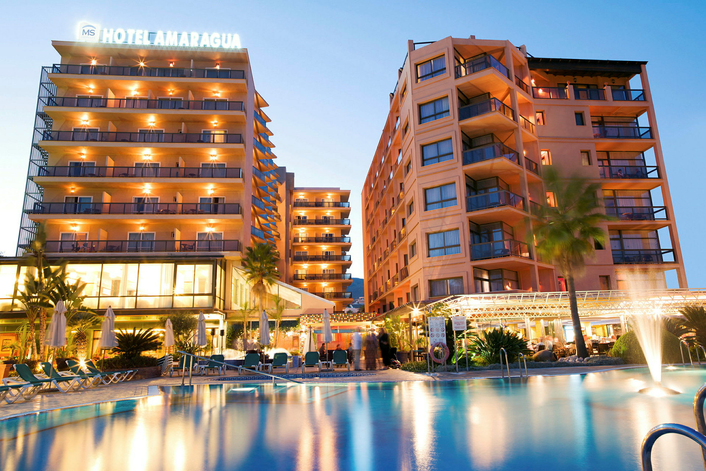 Billede af et hotel med oplyste bygninger og en pool i forgrunden. (AI-genereret tekst)