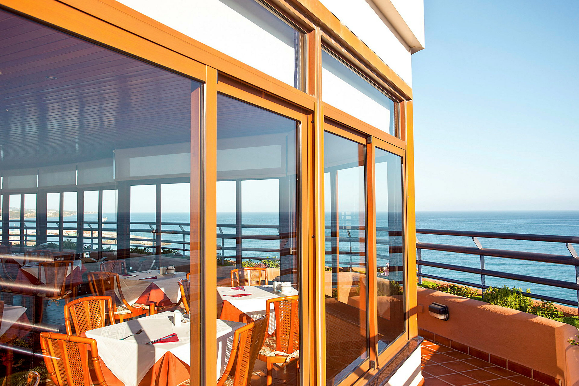 Udsigt over en terrasse med glaspartier, der vender mod havet. Indenfor ses borde med duge og stole. (AI-genereret tekst)