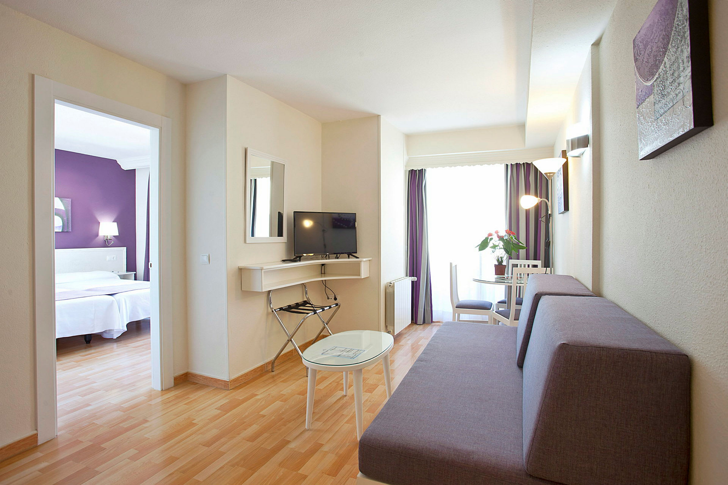 Hotelværelse med opholdsområde, inklusive sofa, tv og bord, samt separat soveværelse synligt gennem en døråbning. (AI-genereret tekst)