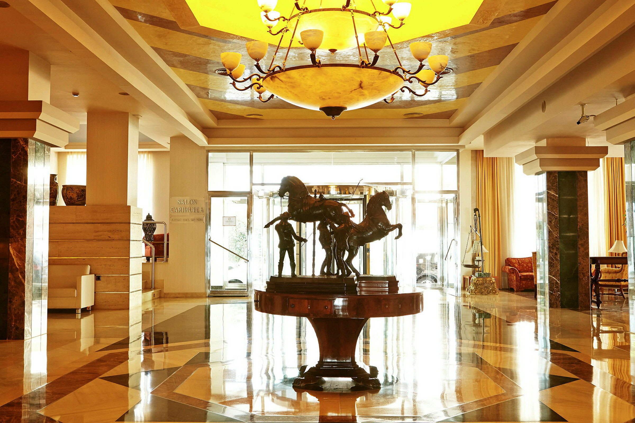 En lys hotellets lobby med et centralt bord, der har en skulptur, der forestiller heste. (AI-genereret tekst)