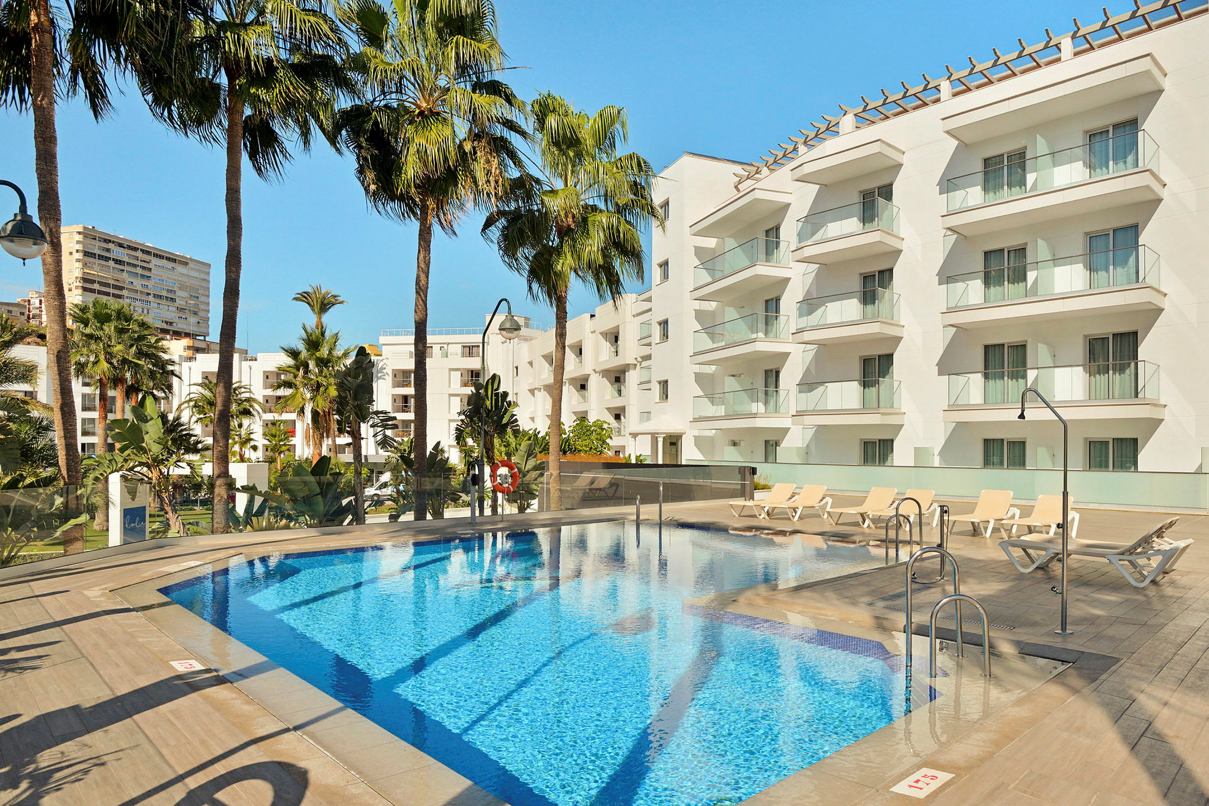 Hotelpool med liggestole og moderne hvide bygninger i baggrunden. (AI-genereret tekst)