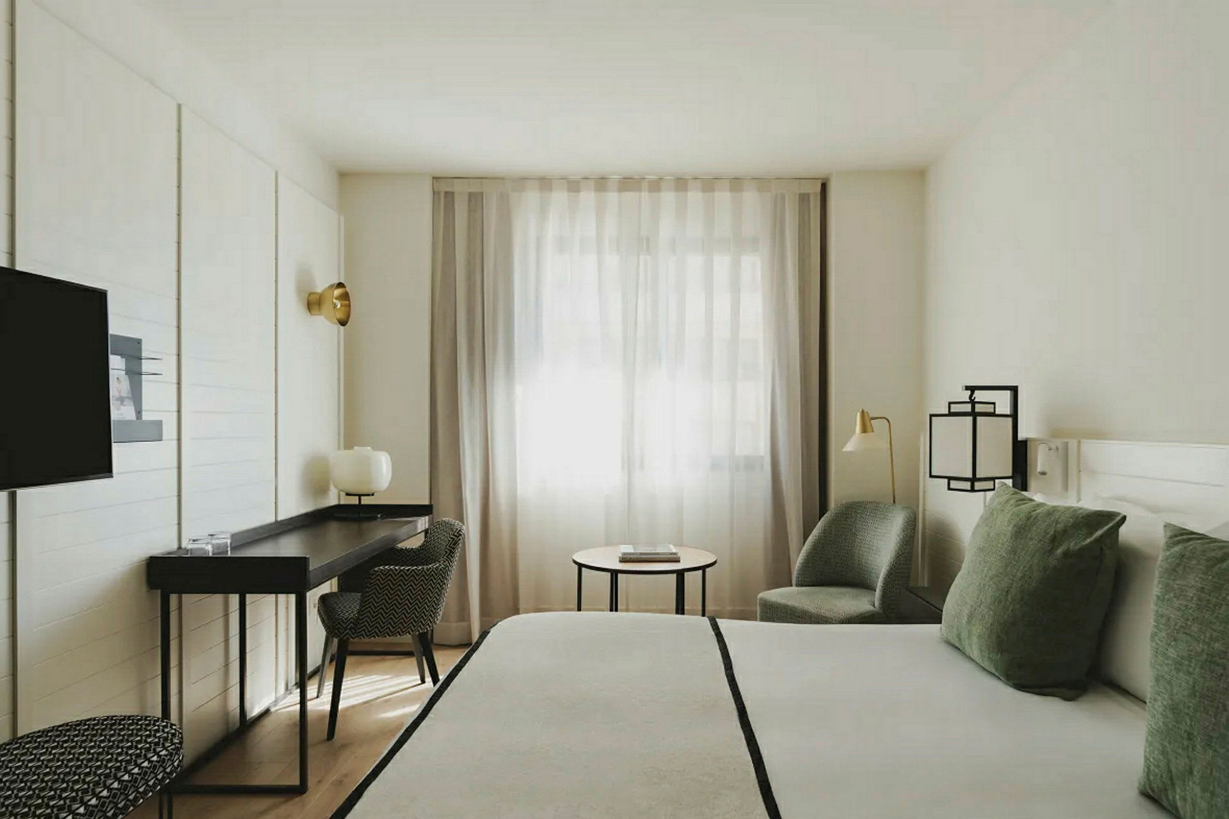 Et lyst hotelværelse med en stor seng, skrivebord, stol og lænestol. Vinduet har lette gardiner. (AI-genereret tekst)
