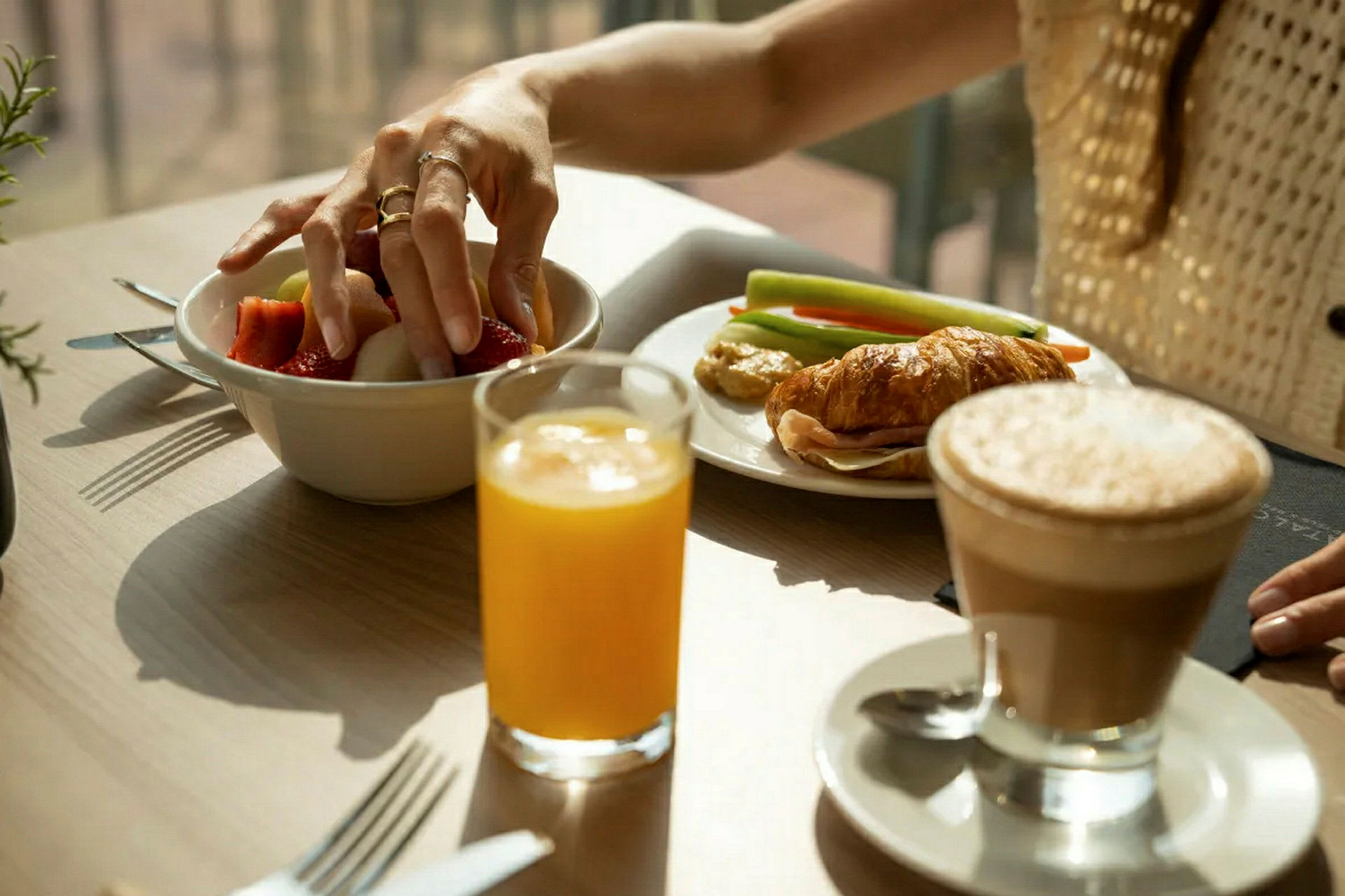 Person tager frugt fra en skål på et bord med croissant, grøntsager, juice og kaffe. (AI-genereret tekst)