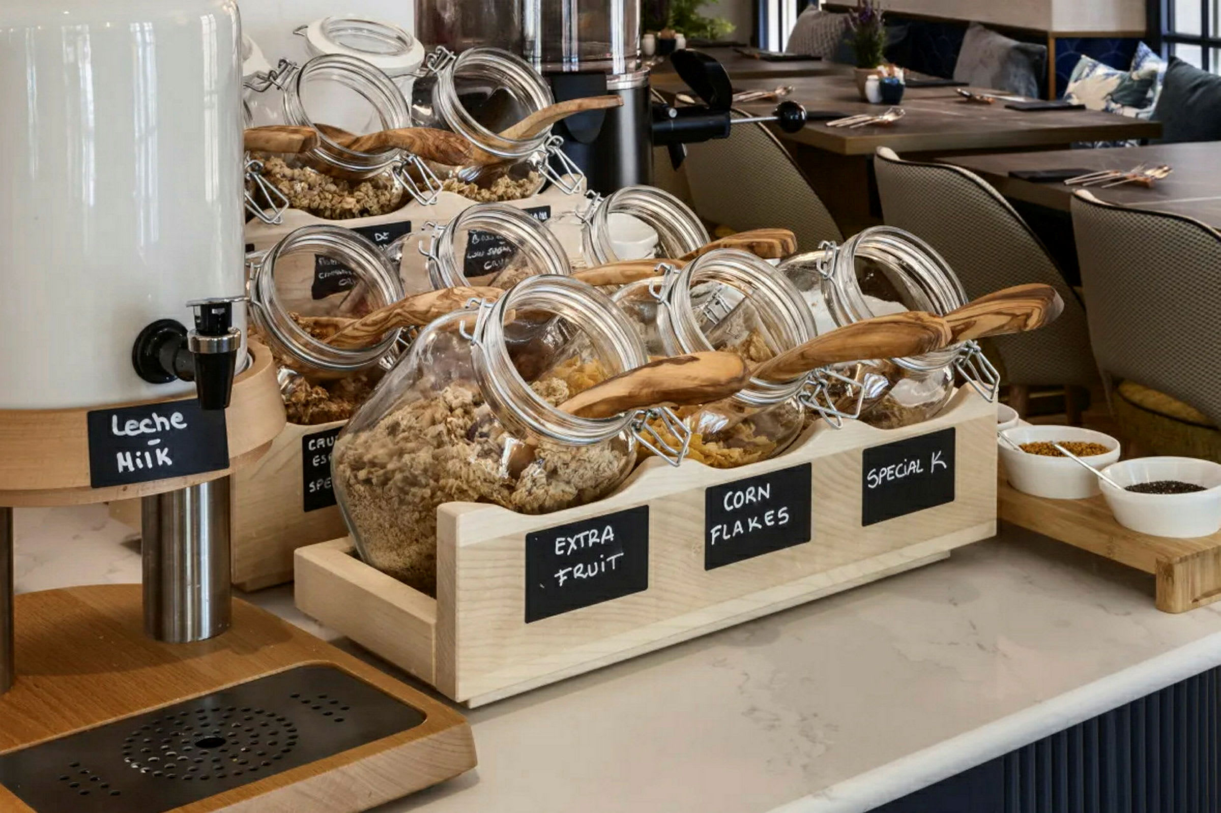 Glaskrukker fyldt med forskellige slags morgenmadsprodukter og müsliblandinger på et hotelbord med mælkebeholder og skåle med toppings. (AI-genereret tekst)