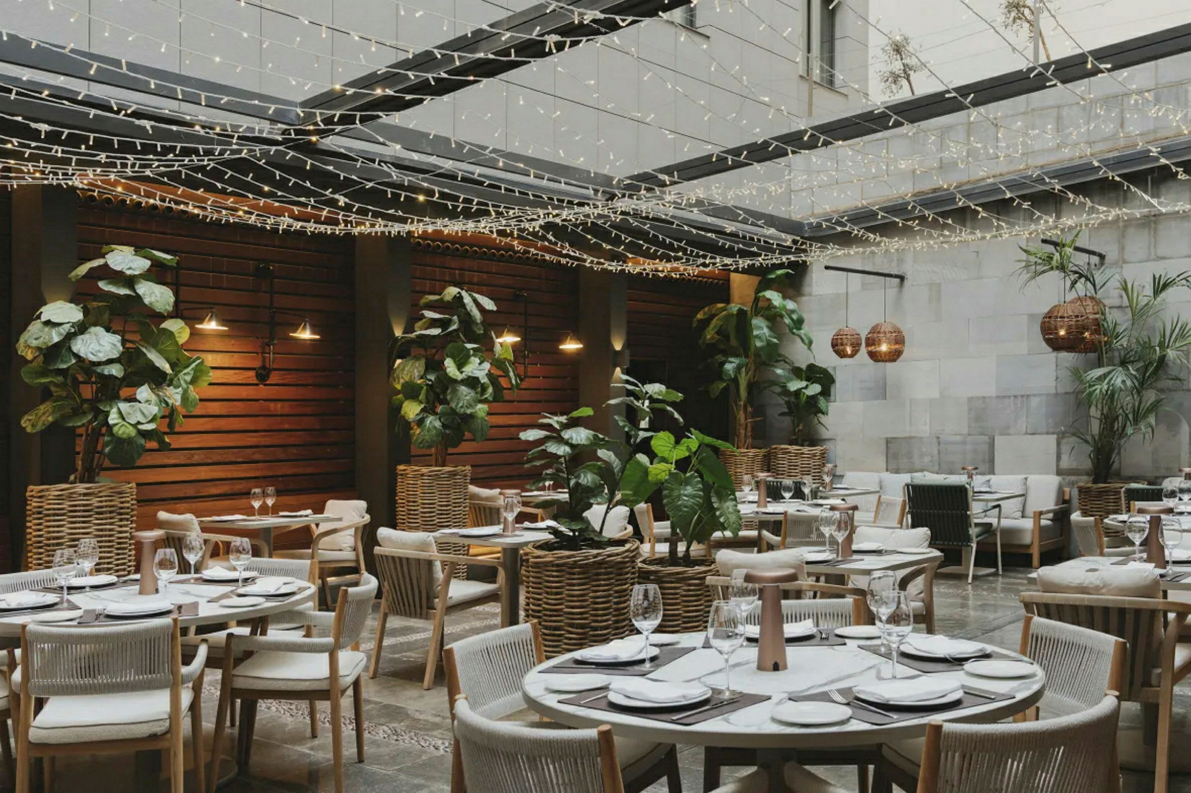 En restaurant med udendørs område, borde dekoreret til måltider omgivet af planter og lyskæder. (AI-genereret tekst)