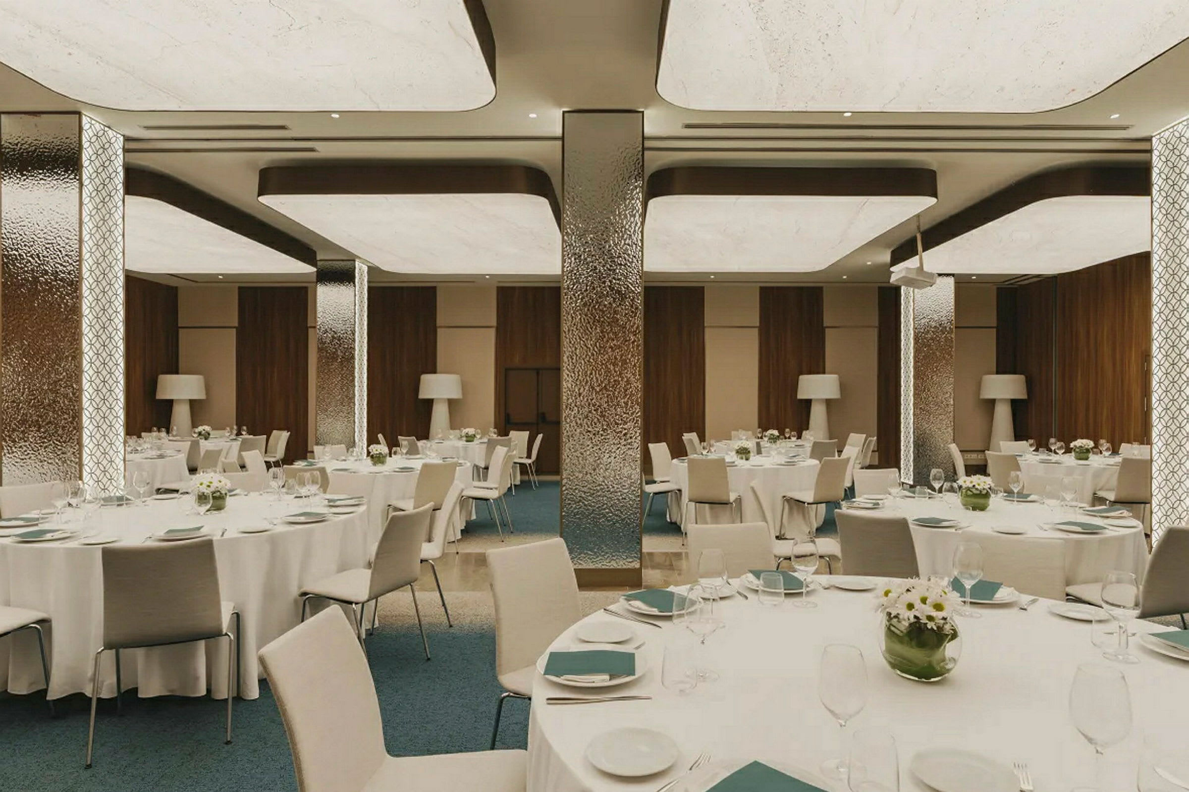En elegant spisesal med dækkede runde borde, dekoreret med blomster og serveringsartikler, omgivet af moderne interiørdetaljer. (AI-genereret tekst)
