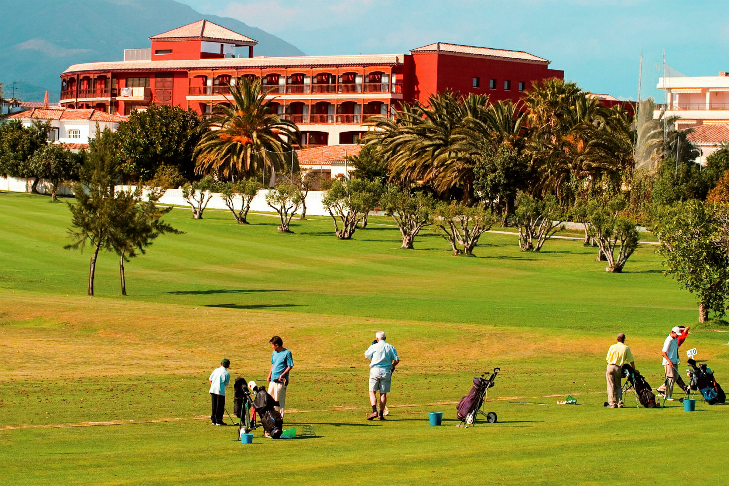Personer spiller golf på en grøn golfbane foran et rødt hotel. (AI-genereret tekst)