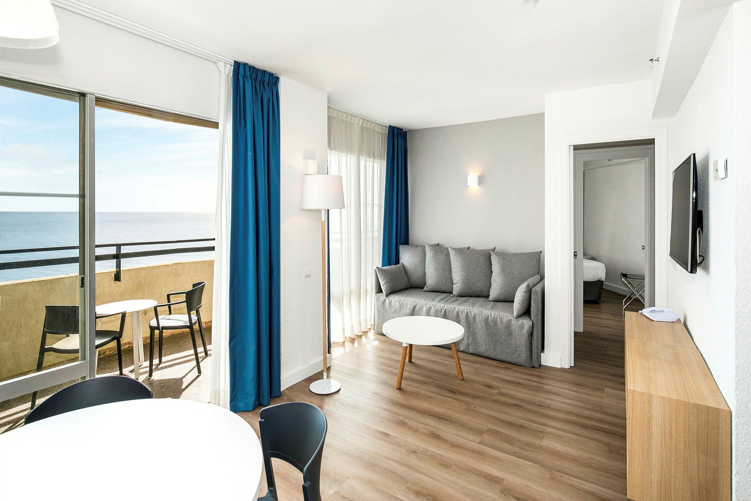 En stue med en grå sofa, et rundt sofabord og et altanområde med havudsigt. (AI-genereret tekst)