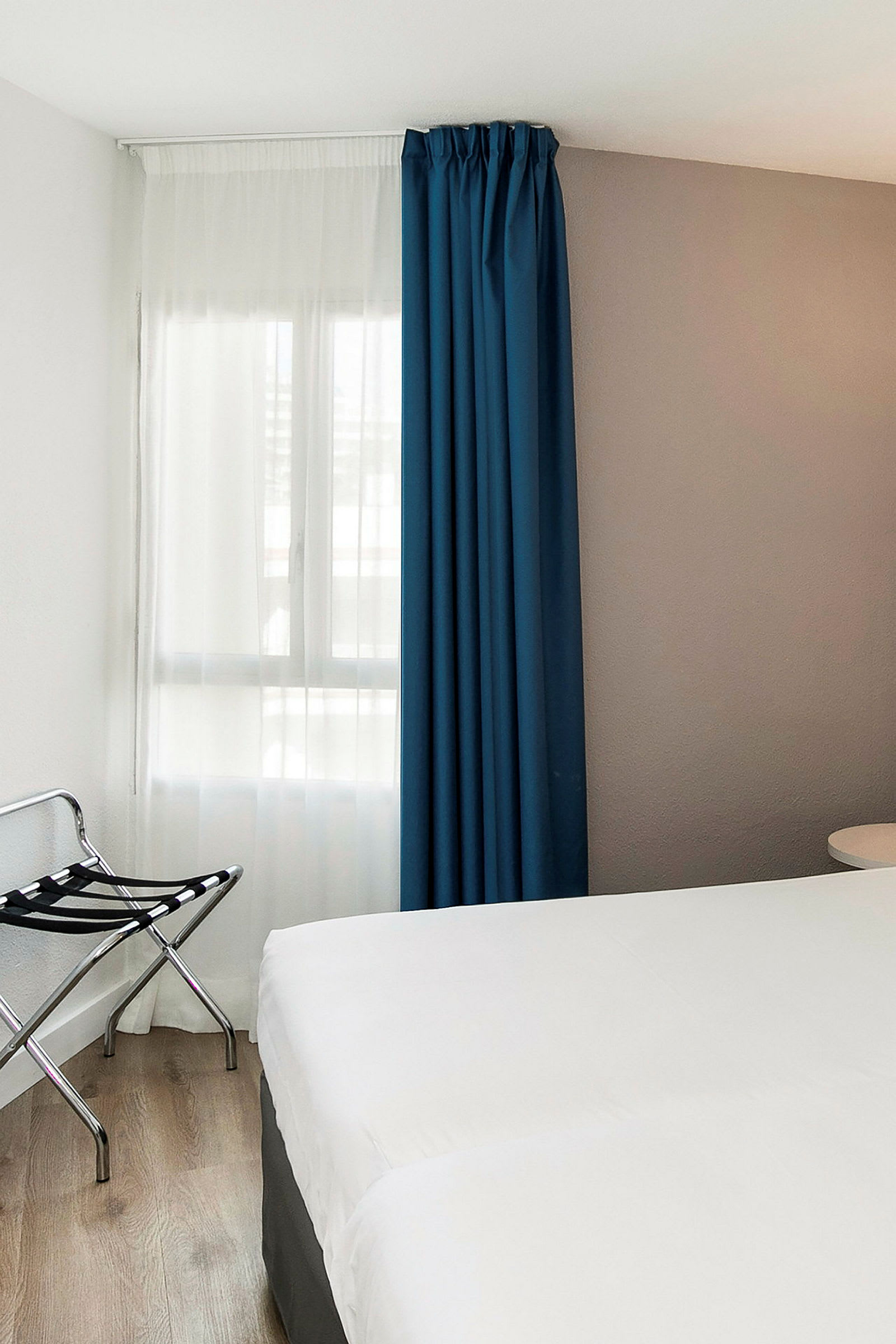 Et hotelværelse med et hvidt sengetæppe, et blå gardin og en bagageholder ved siden af. (AI-genereret tekst)