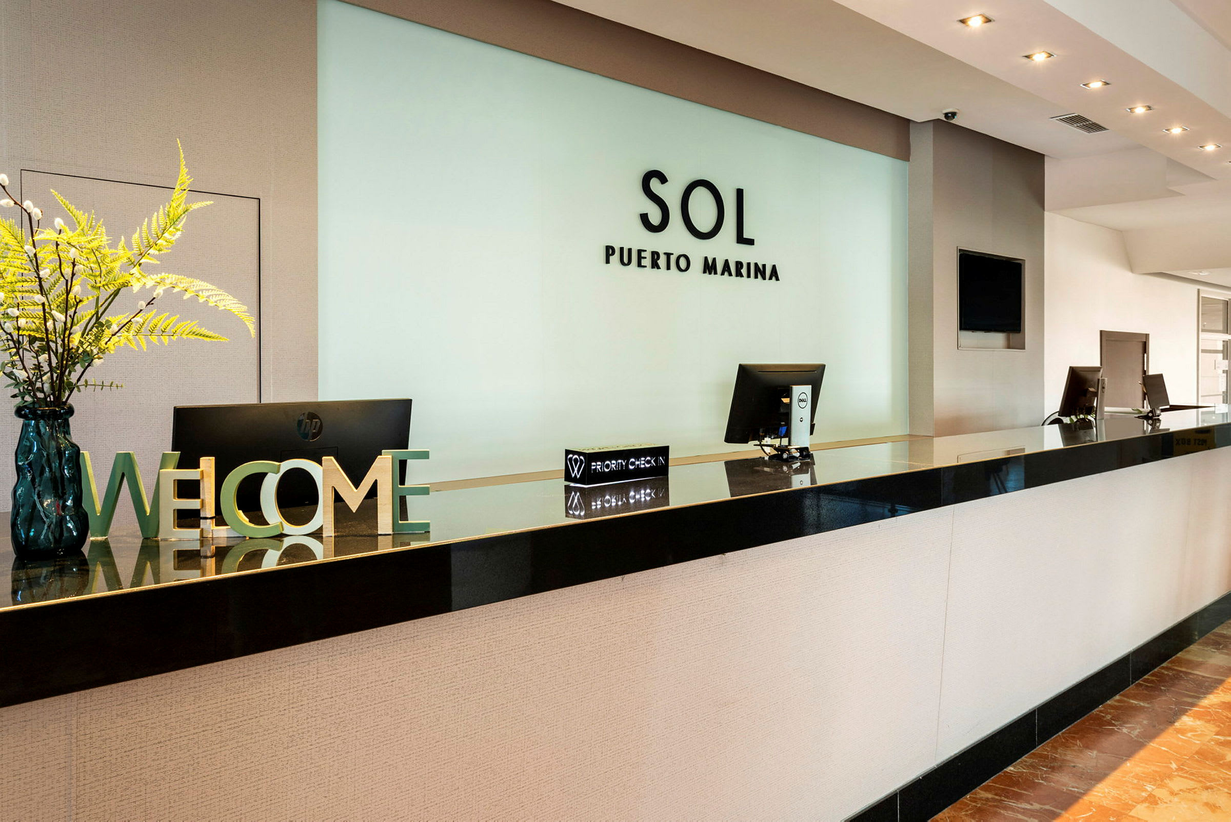 Receptionen på Sol Puerto Marina med computerskærme og dekorationer, herunder et velkomstskilt og blomsterarrangement. (AI-genereret tekst)