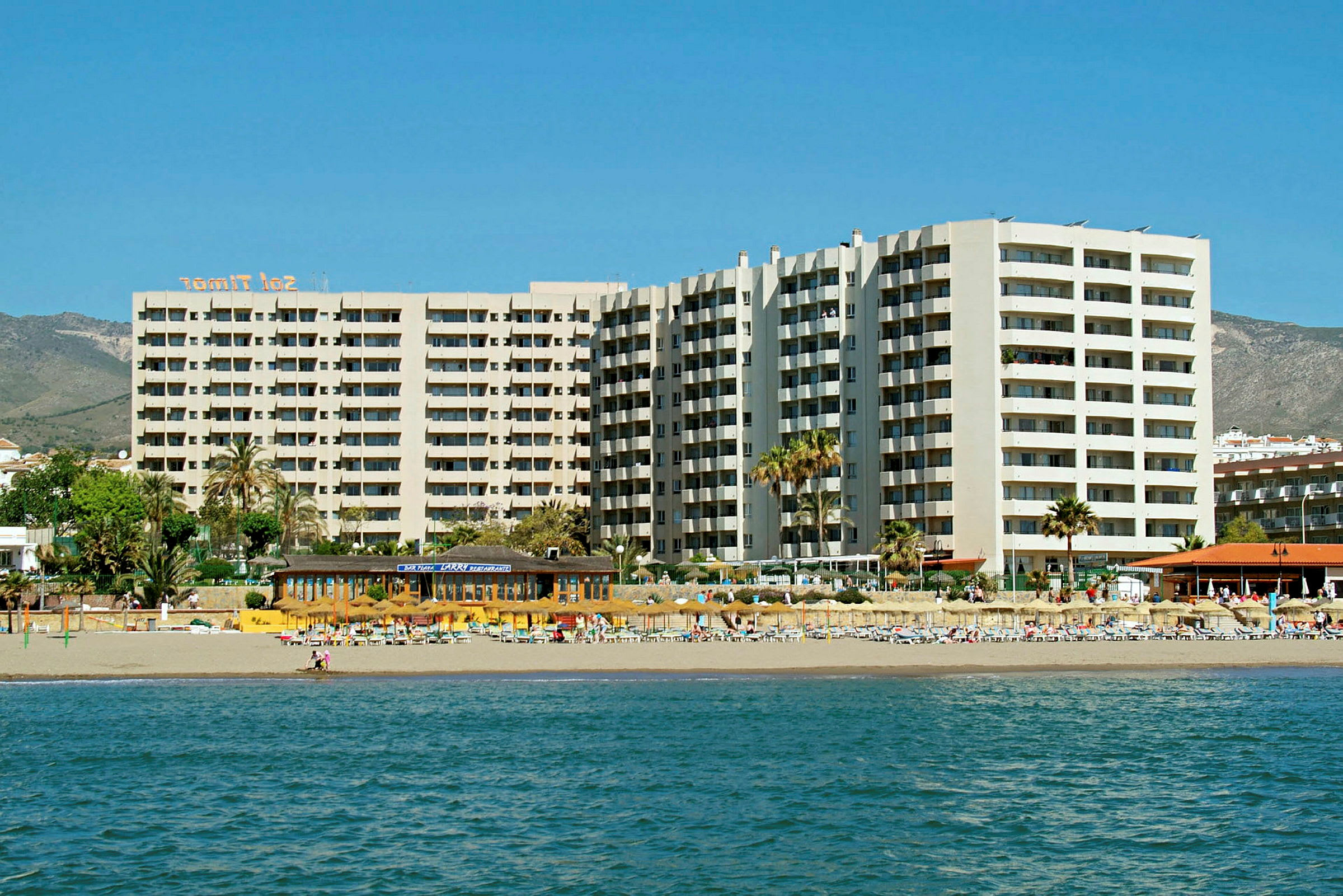 Strandhotel med høje bygninger og liggestole ved vandet. (AI-genereret tekst)