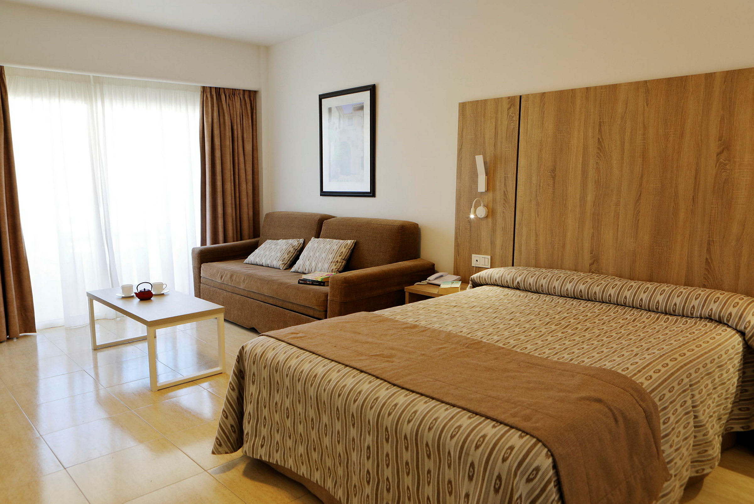 Hotelværelse med en brun sofa, hvide gardiner, et bord og en seng. (AI-genereret tekst)