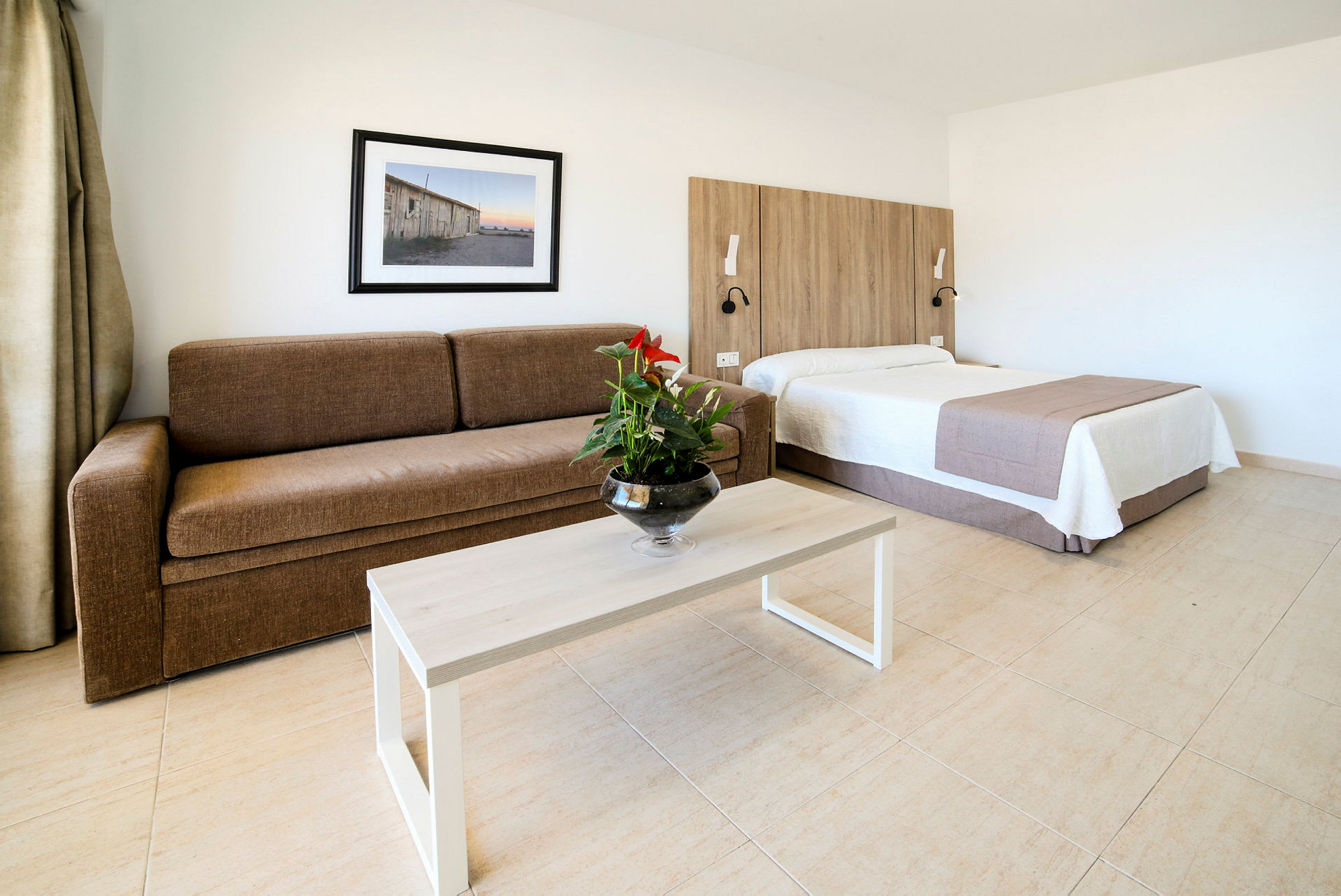 Et hotelværelse med en brun sofa, et bord med en blomsterkrukke, og en seng med træpanel bagved. (AI-genereret tekst)