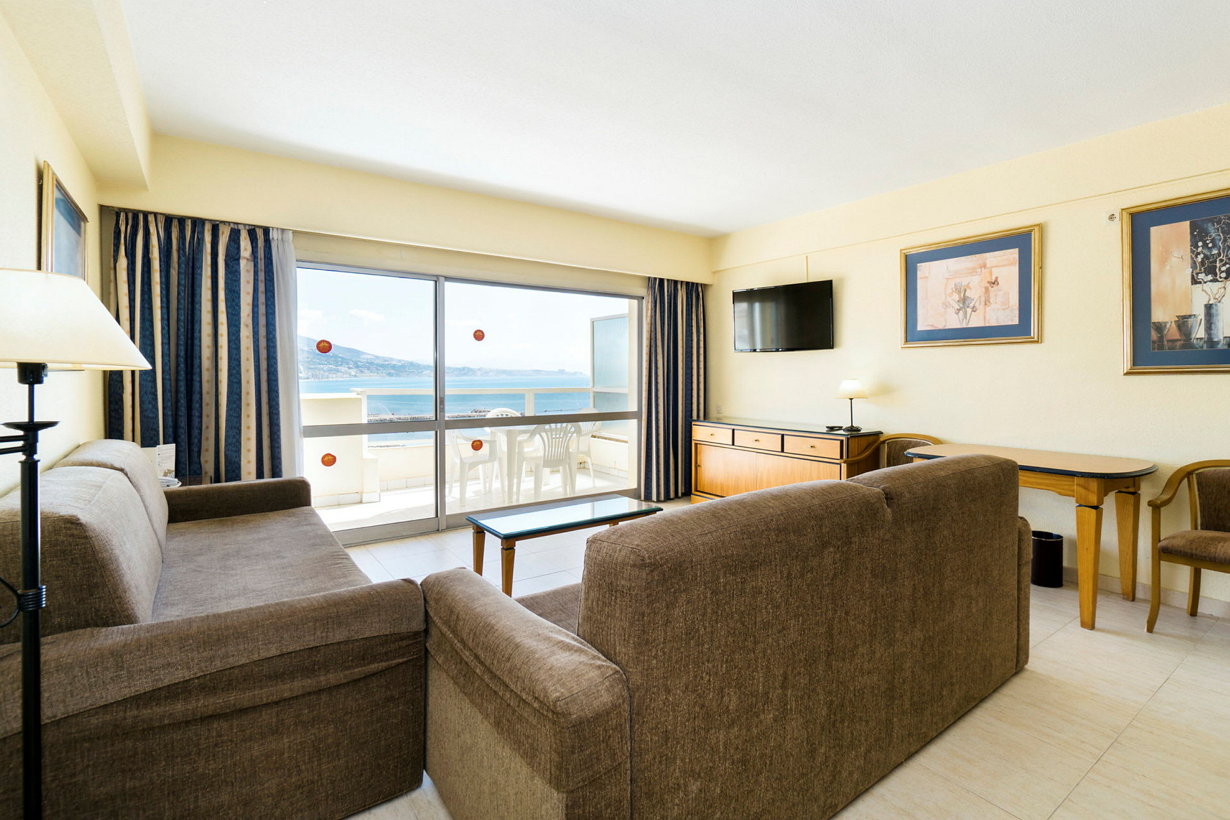 Stue med sofaer, bord, TV og udgang til balkon med udsigt over havet. (AI-genereret tekst)