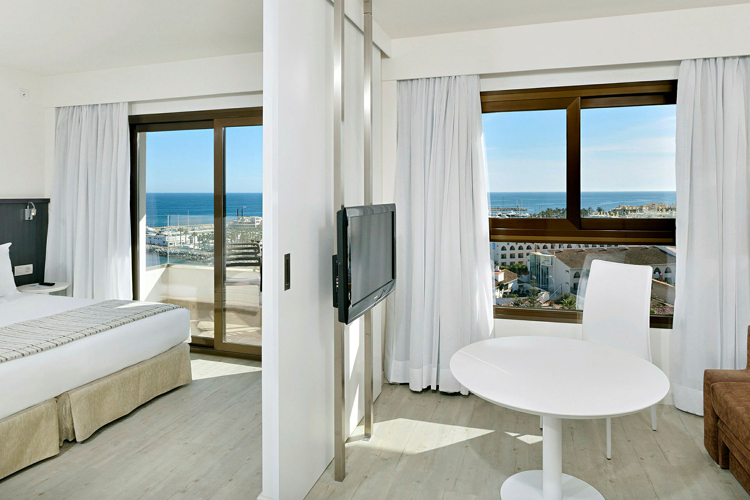 Hotelværelse med to vinduer der vender ud mod havet, en seng, et TV og et hvidt bord med en stol. (AI-genereret tekst)
