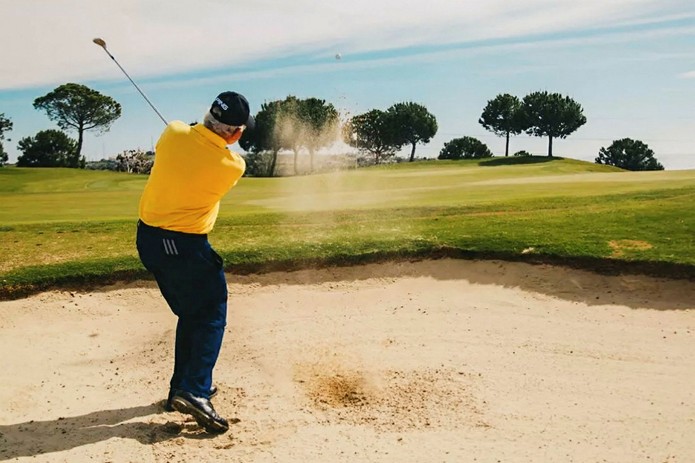 En person slår en golfbold fra en sandbunker på en golfbane omgivet af grønne træer. (AI-genereret tekst)