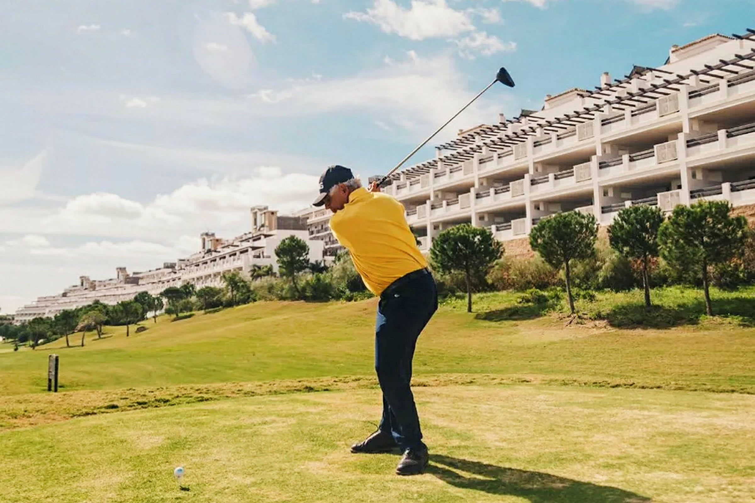 En person i gul trøje spiller golf på en grøn bane med en bygning og træer i baggrunden. (AI-genereret tekst)