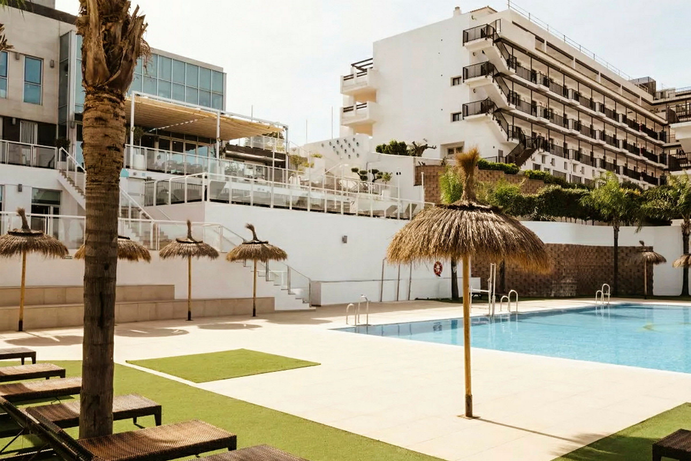En pool med parasoller og liggestole foran et fleretasjes hotel. (AI-genereret tekst)