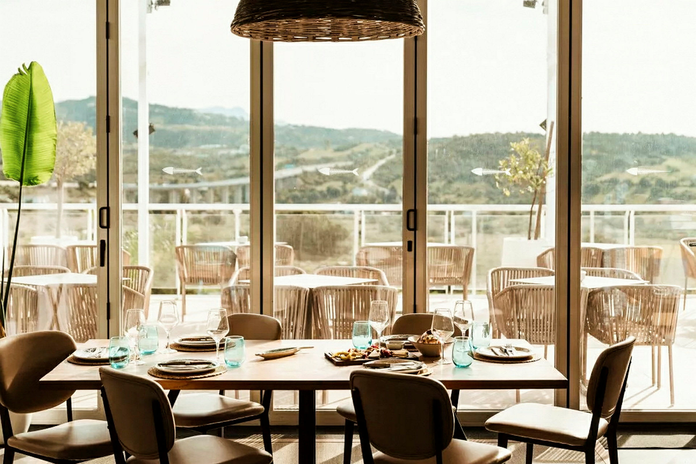 En restaurant med et dækket bord og udsigt over en terrasse med stole og bjerge i det fjerne. (AI-genereret tekst)