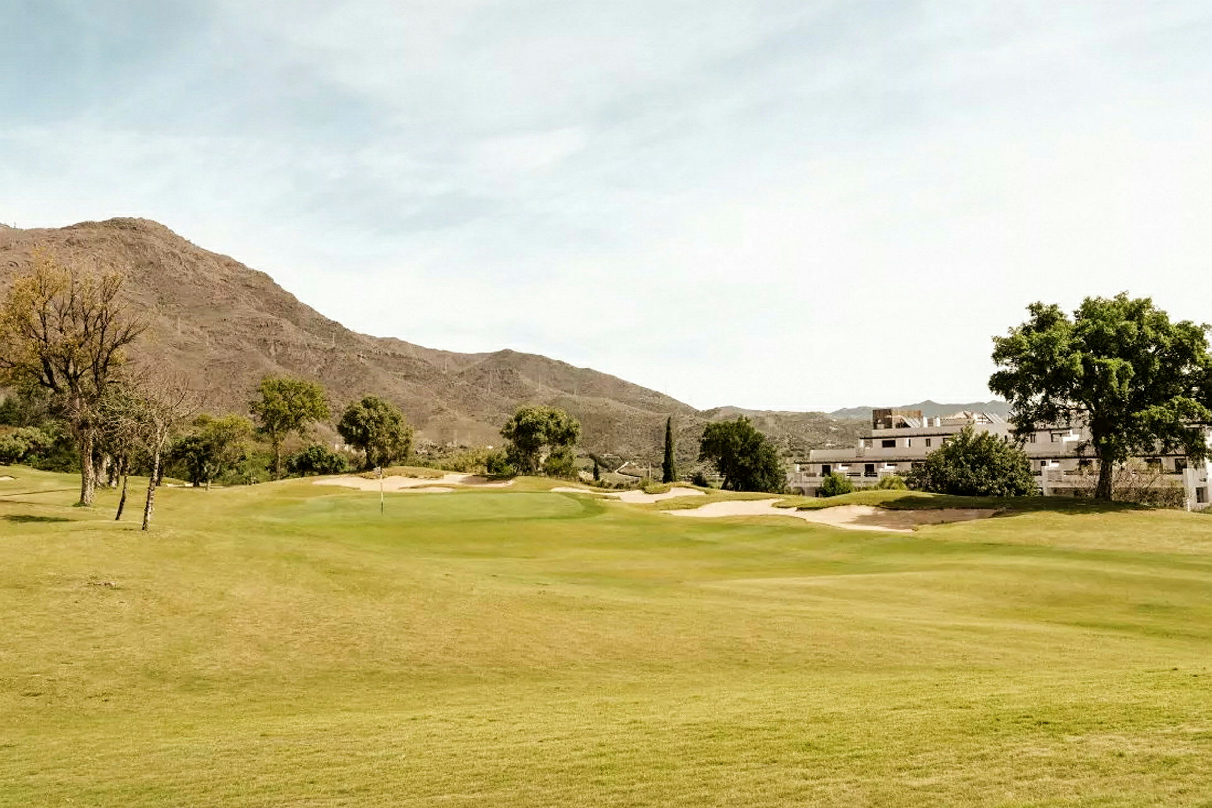 En golfbane omgivet af grøn vegetation, med bygninger og bjerge i baggrunden. (AI-genereret tekst)