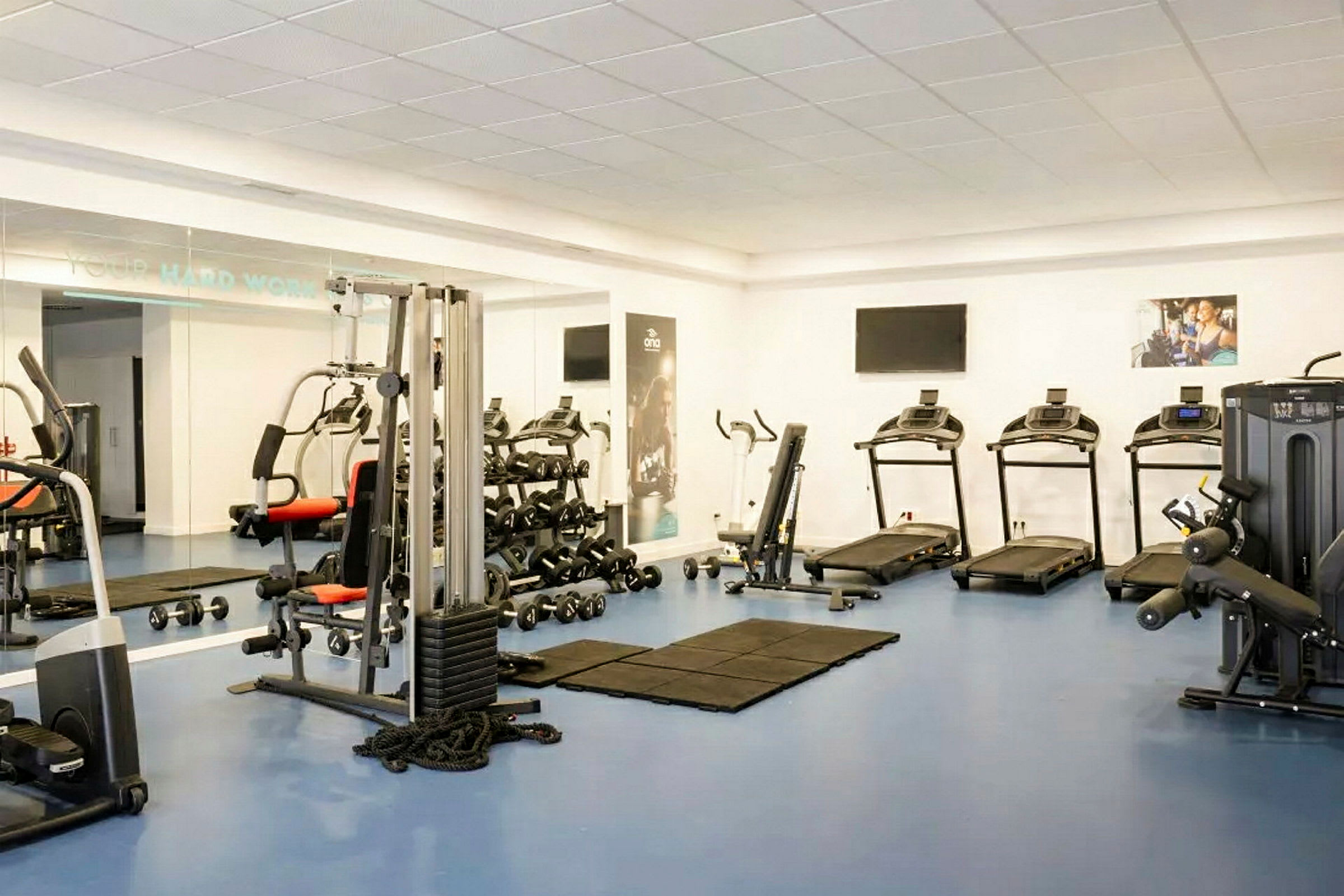 Et fitnesscenter med udstyr som løbebånd, træningsmaskiner og håndvægte. Spejle dækker den ene væg. (AI-genereret tekst)