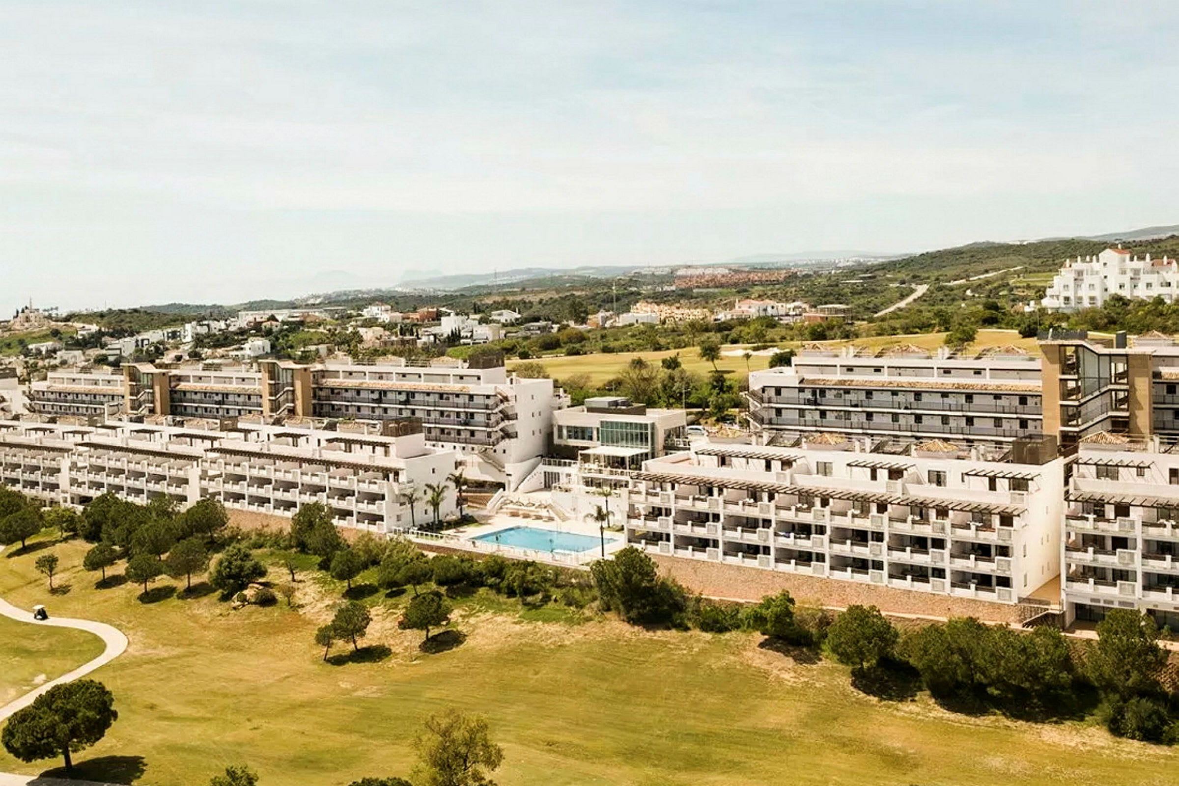 Luftfoto af et hotelområde med flere etagebygninger, omgivet af grønne områder og en pool. (AI-genereret tekst)