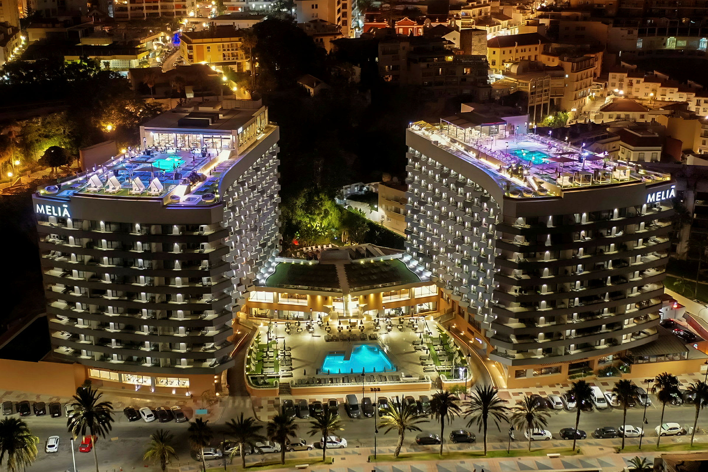 Et hotelkompleks om aftenen med to højhuse, en oplyst pool og terrasser. (AI-genereret tekst)