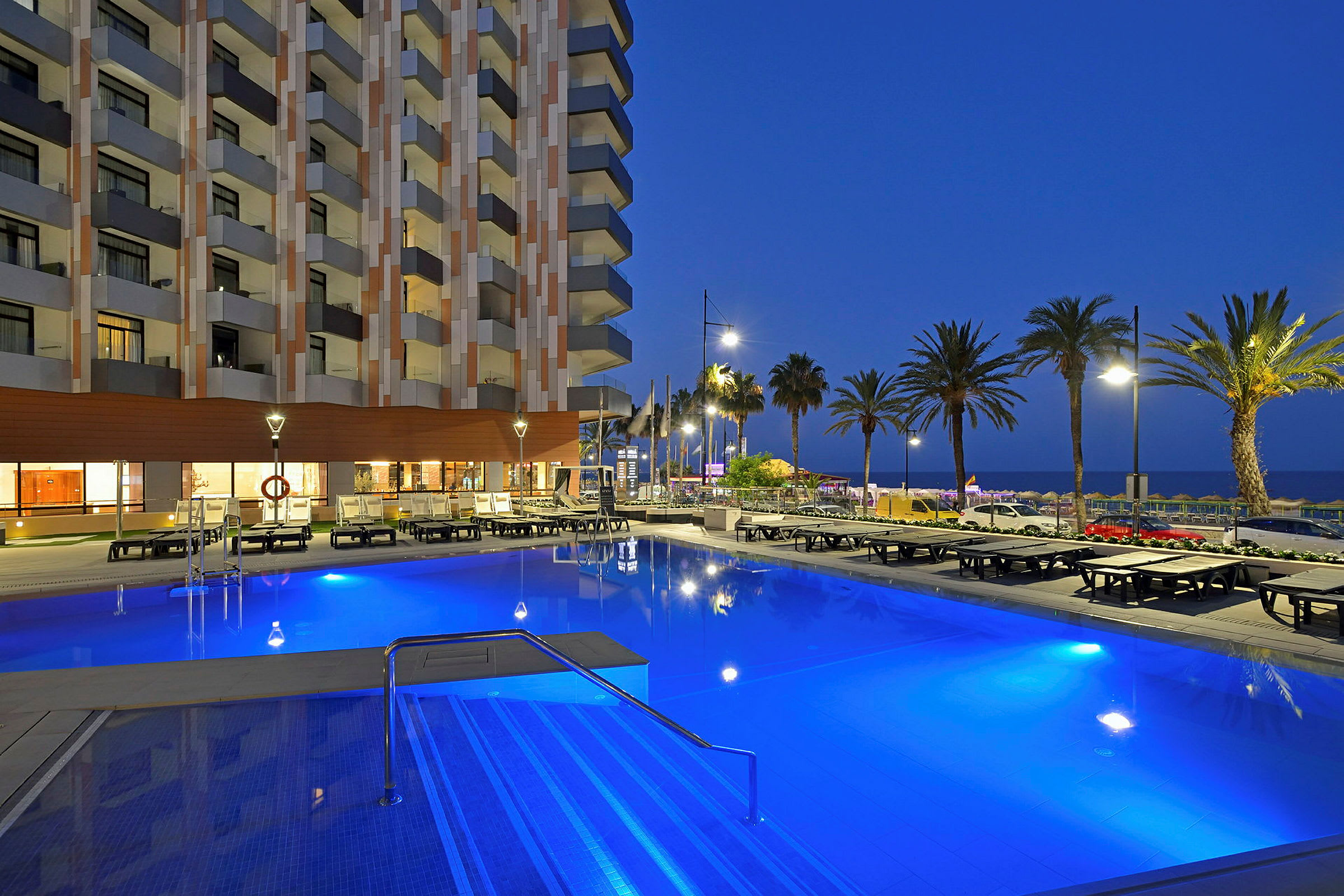 Udendørs pool ved et hotel omgivet af liggestole. Baggrunden viser palmer og havet om aftenen. (AI-genereret tekst)