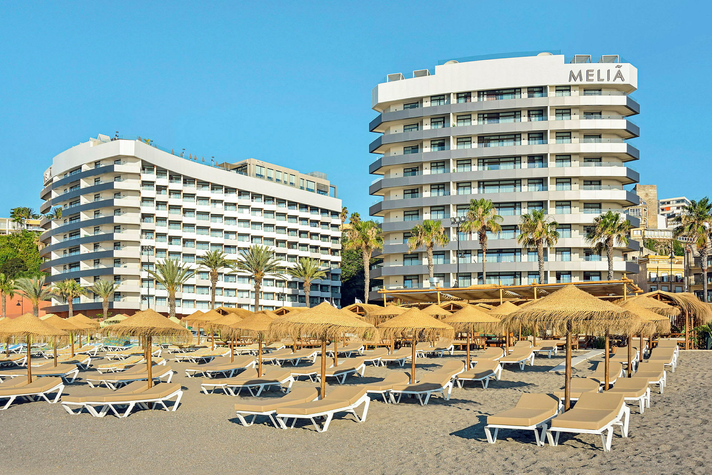 Billede af to hotelbygninger med altaner. Foran er der parasoller og solsenge på en strand. (AI-genereret tekst)
