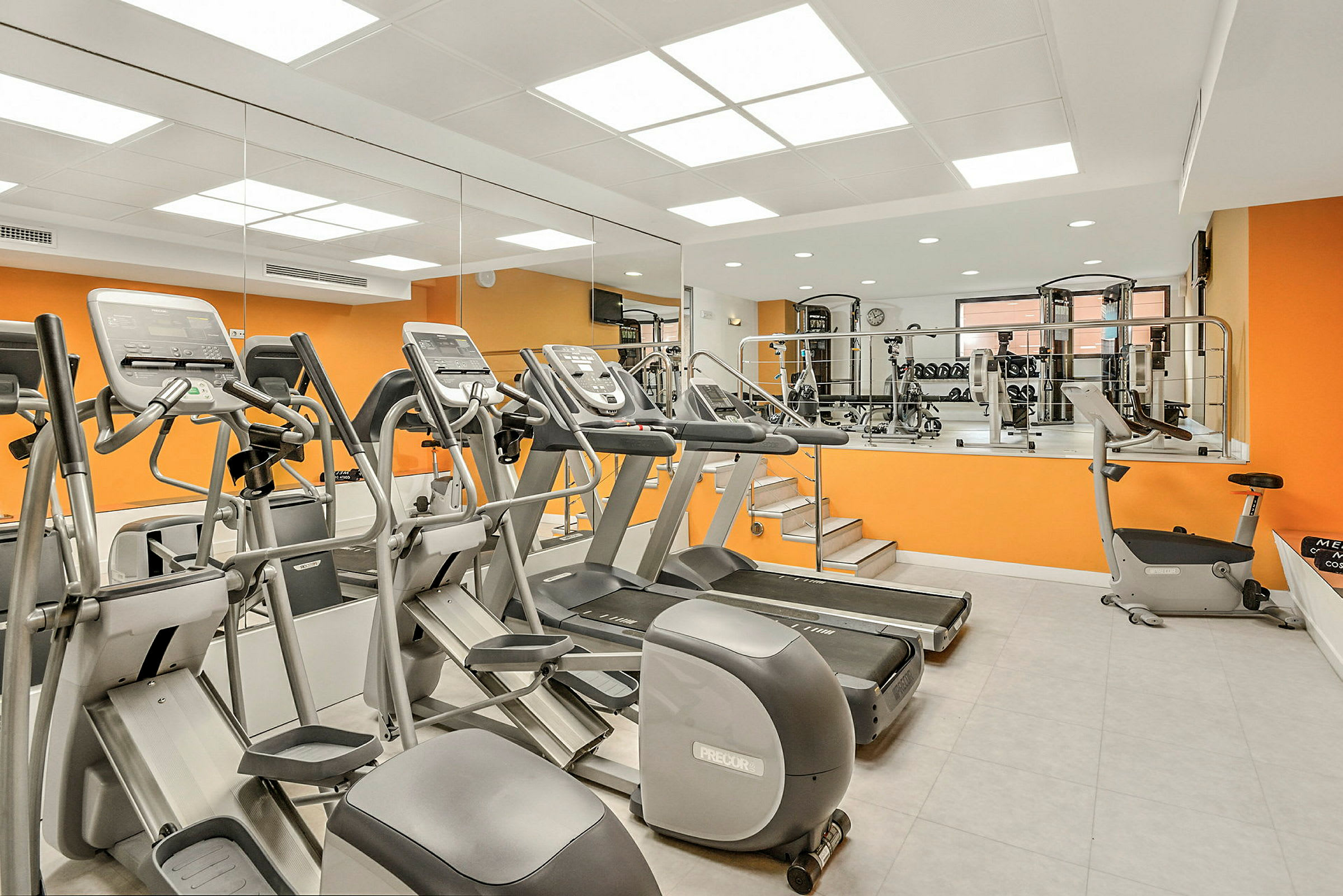 Et fitnesscenter med træningsmaskiner, løbebånd og en orange væg. (AI-genereret tekst)