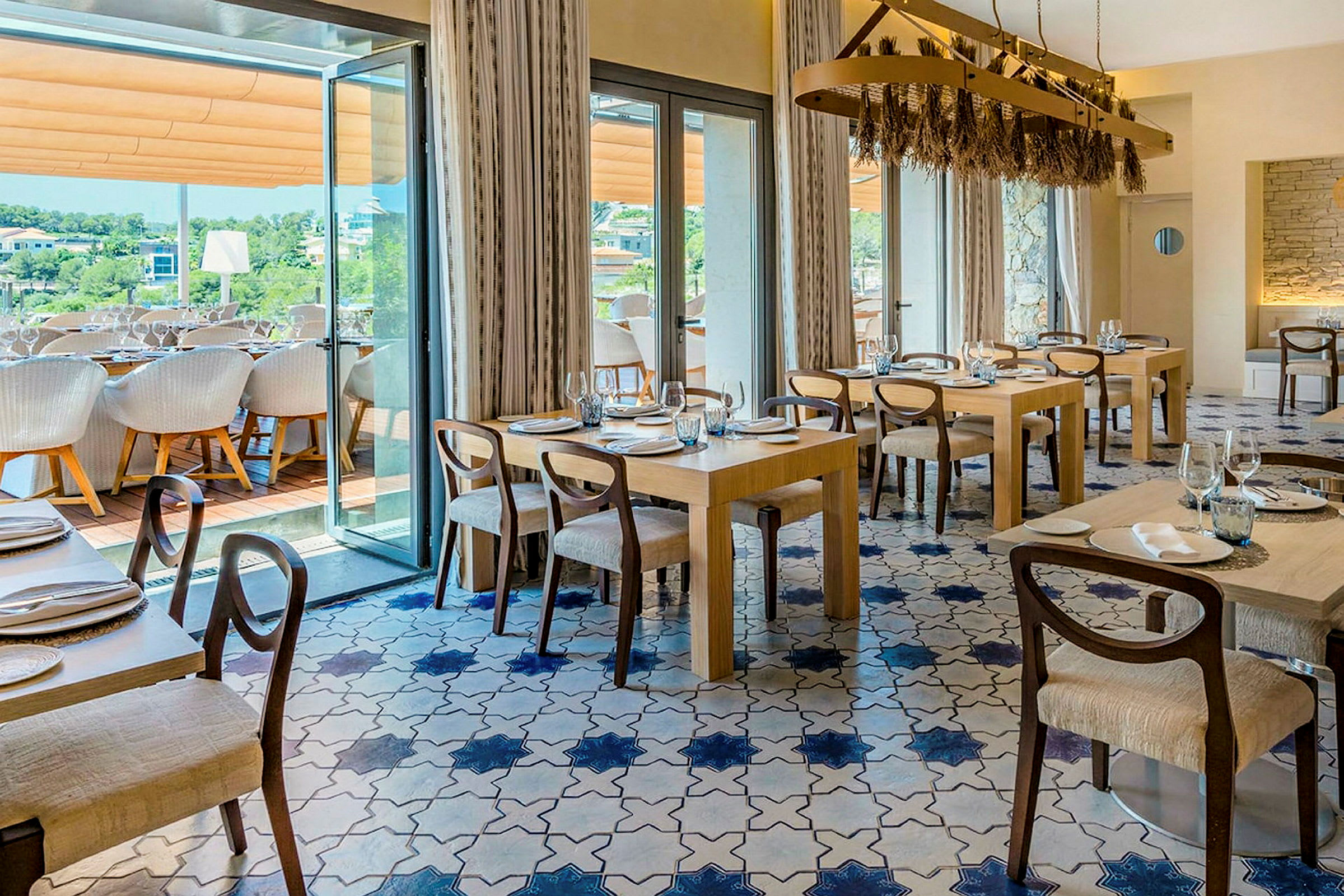 En elegant restaurant med flere borde og stole, blå og hvide fliser på gulvet samt store vinduer, der fører ud til en terrasse. (AI-genereret tekst)