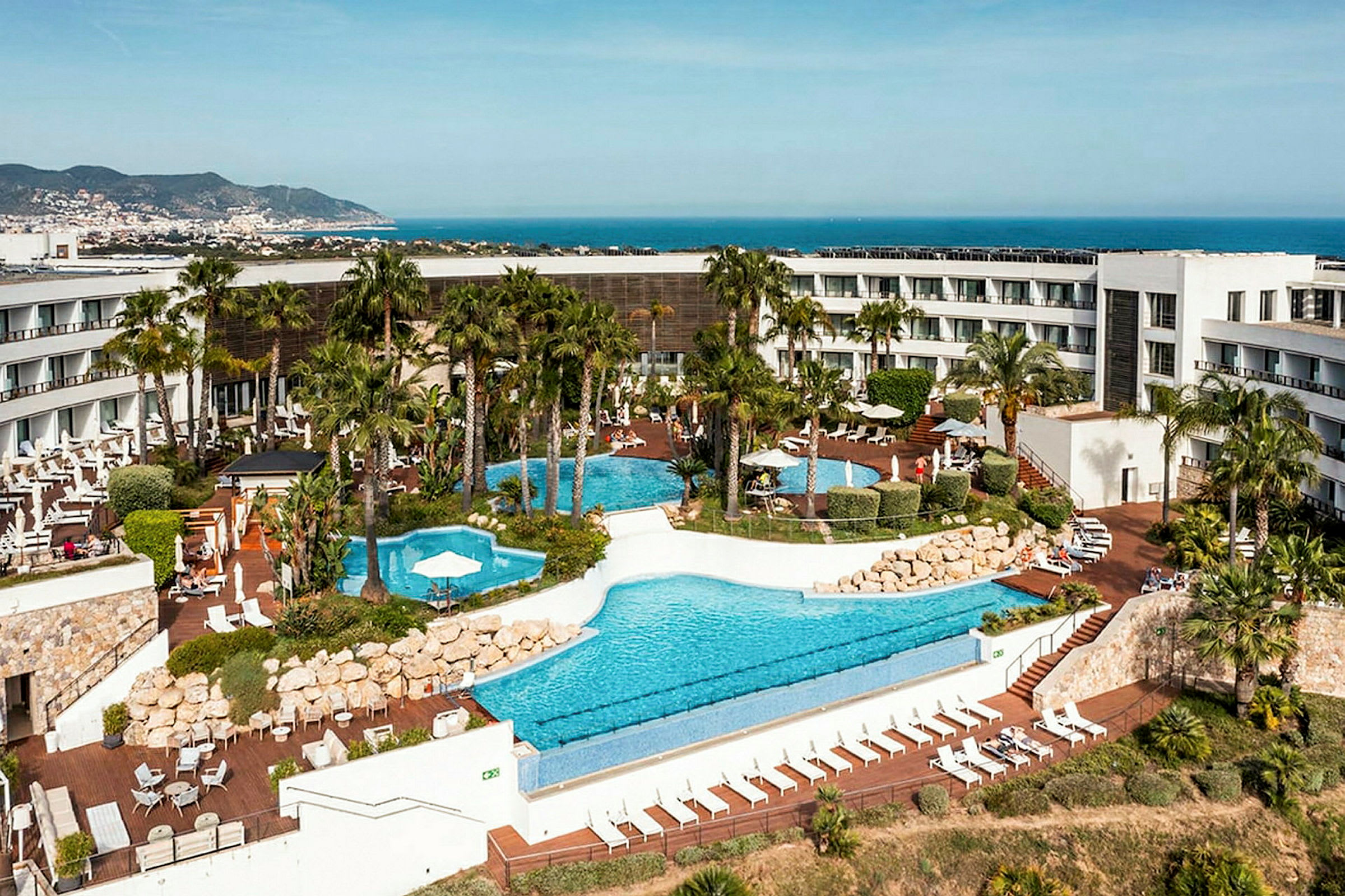 Et hotel med poolområde og omgivende have, med havet og en by i baggrunden. (AI-genereret tekst)