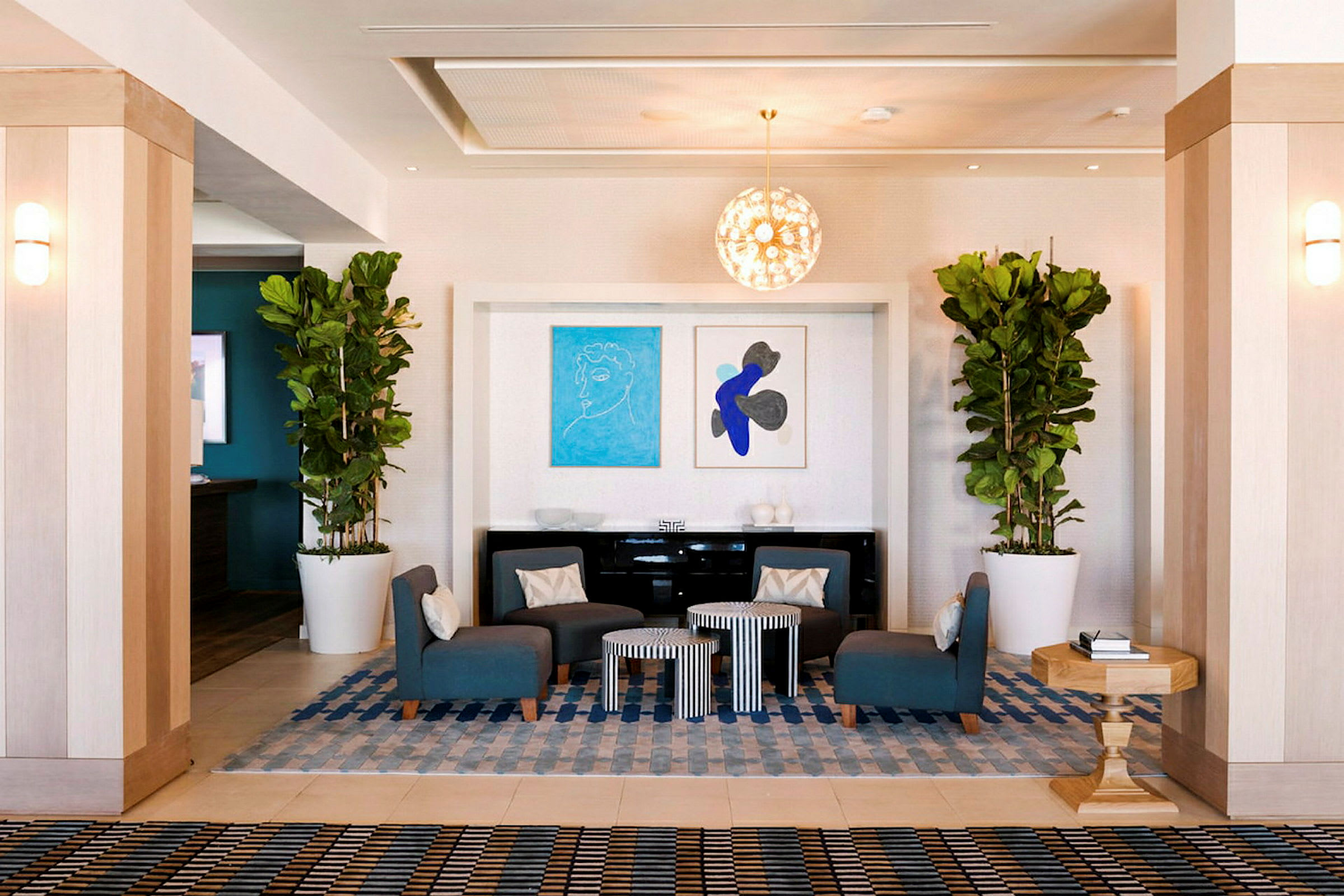 En hotellobby med siddepladser i form af blå stole omkring små borde. Planter i store hvide potter og kunstværker på væggen dekorerer rummet. (AI-genereret tekst)