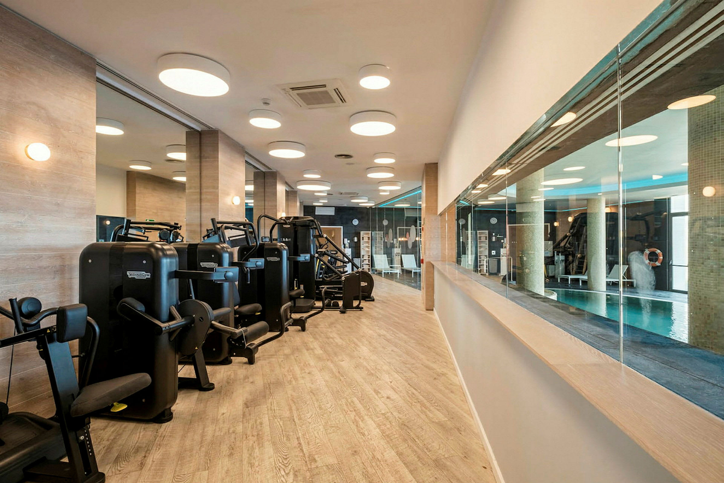 Et fitnesscenter med træningsmaskiner og spejle på væggene samt en glasvæg med udsigt til en indendørs pool. (AI-genereret tekst)