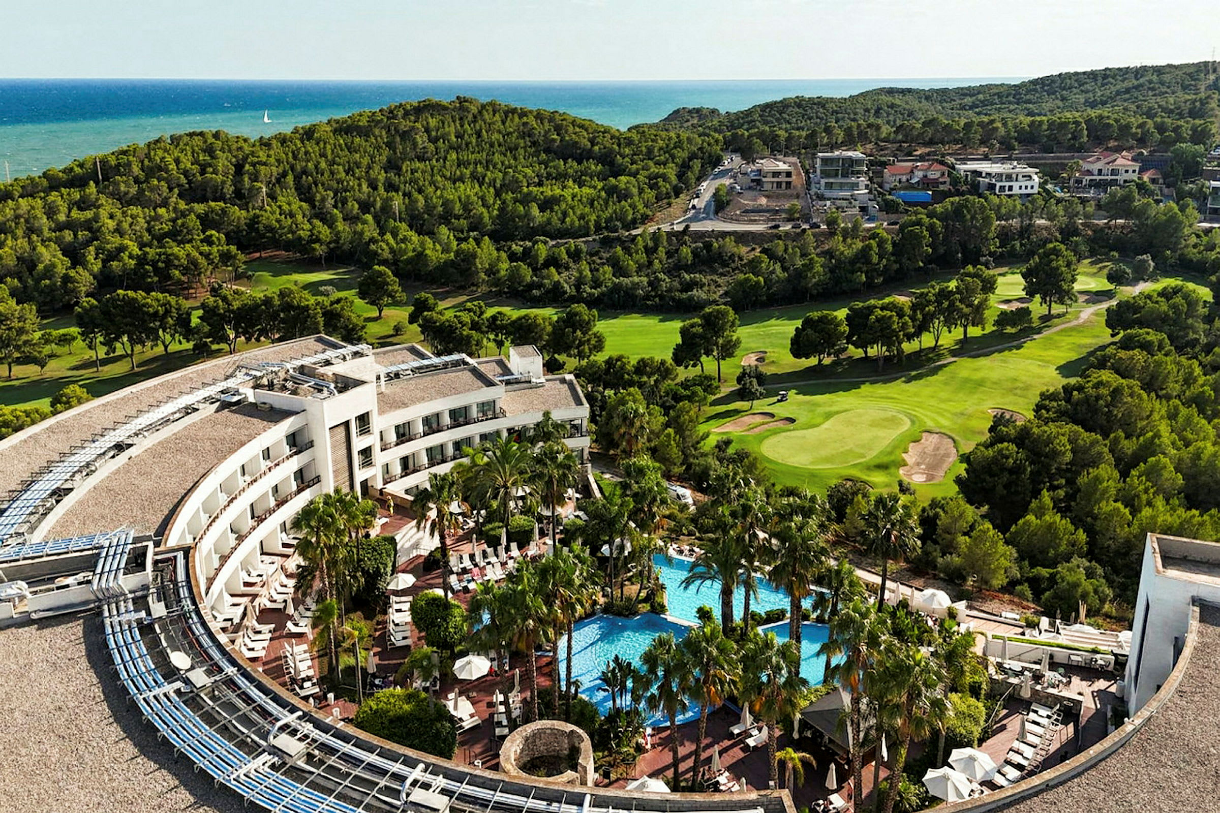 Billede af en hotelbygning med pool og omkringliggende palmer, omgivet af græsplæner og skov med havet i baggrunden. (AI-genereret tekst)