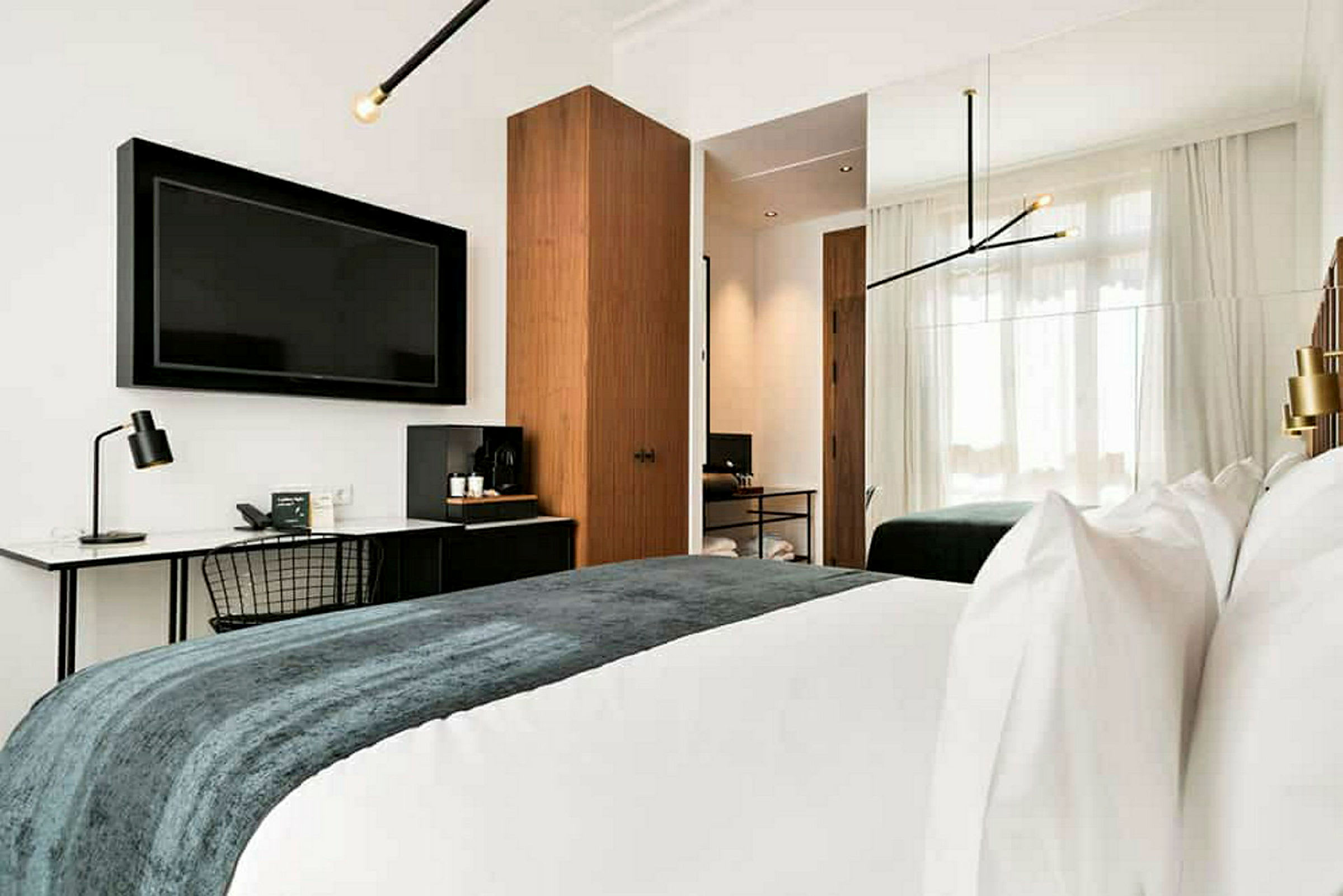 Billede af et moderne hotelværelse med et stort TV på væggen, skrivebord med lampe, en dobbeltseng med hvidt sengetøj og et tæppe i mørke farver. Værelset har også et skab og store vinduer med lyse gardiner. (AI-genereret tekst)