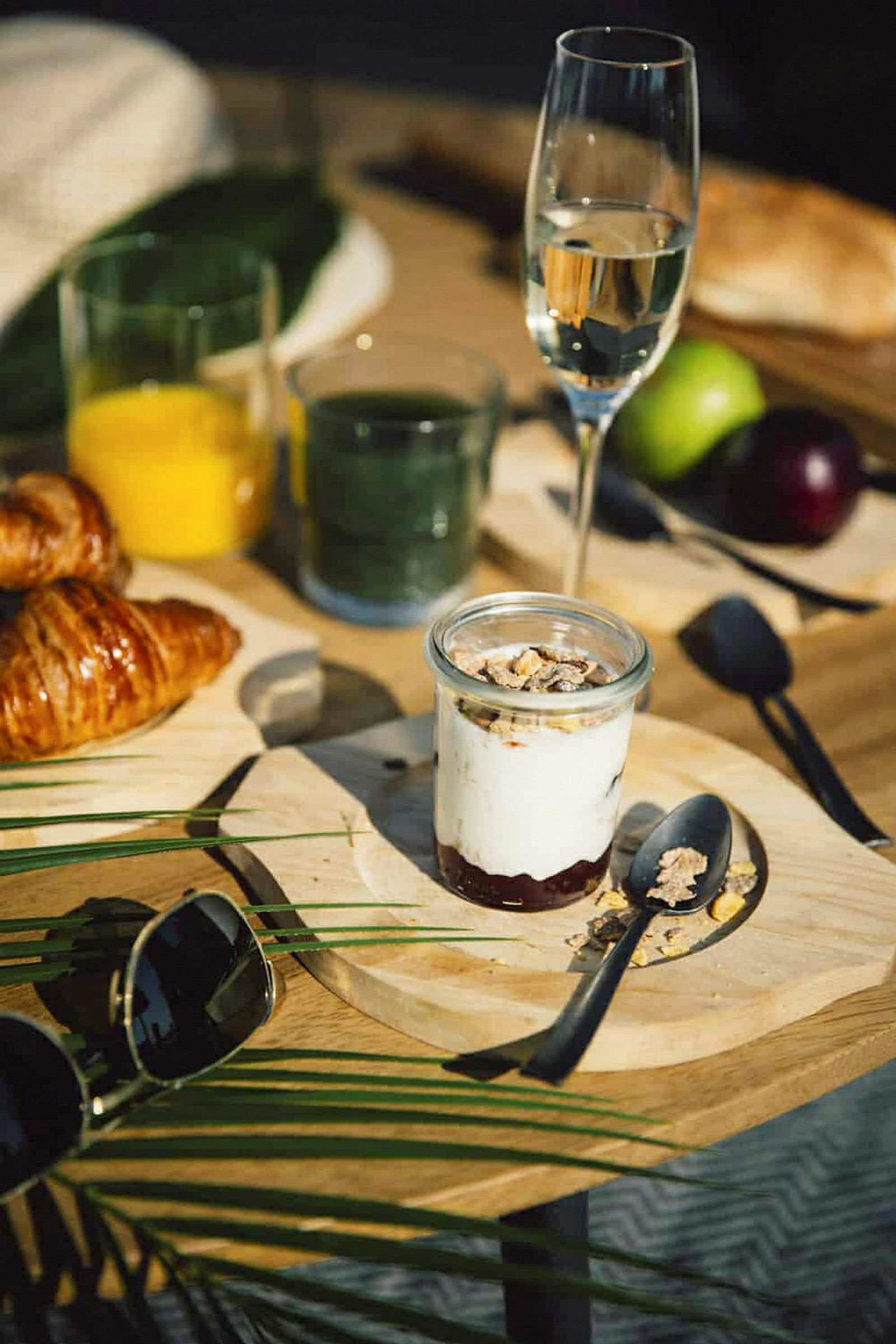 Et morgenbord med glas med drikke, en croissant, en tallerken med yoghurt og granola samt andre detaljer som briller og frugt. (AI-genereret tekst)