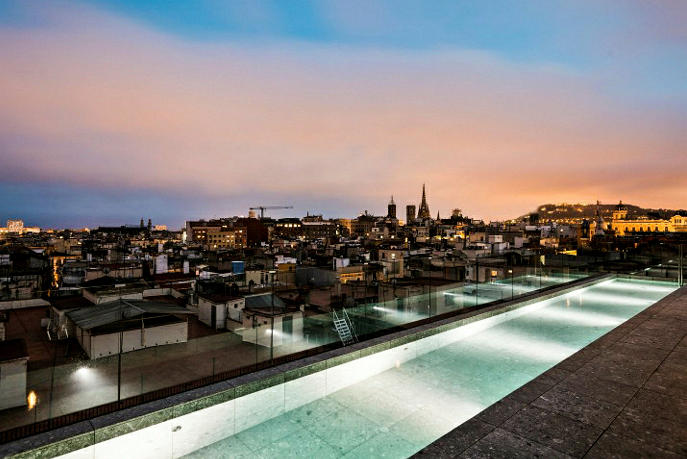 Udsigt fra en tagterrasse med en lang pool over en by i skumringen. (AI-genereret tekst)