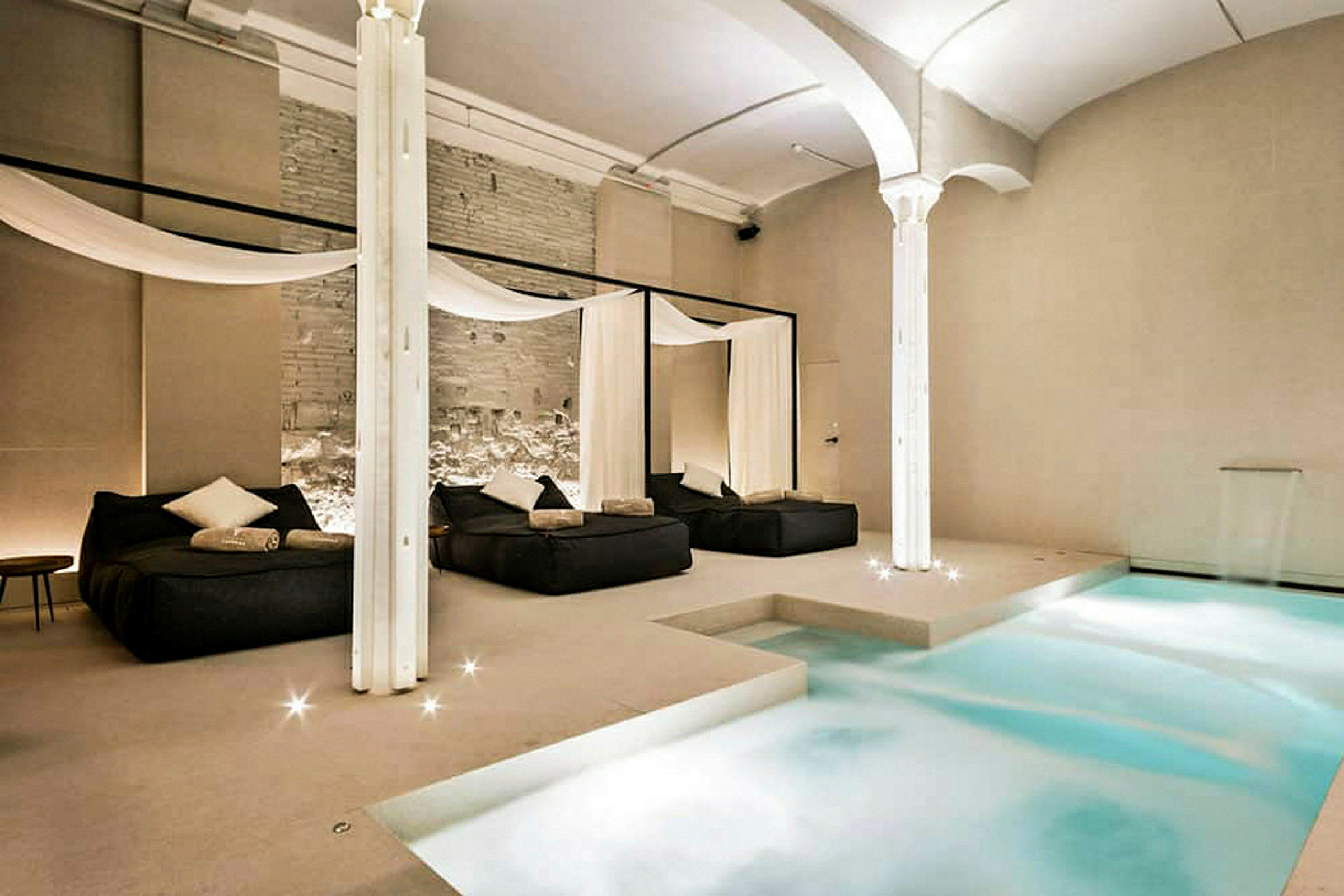 Et elegant indendørs spa med en pool og afslapningsområder med senge og dekorative gardiner. (AI-genereret tekst)