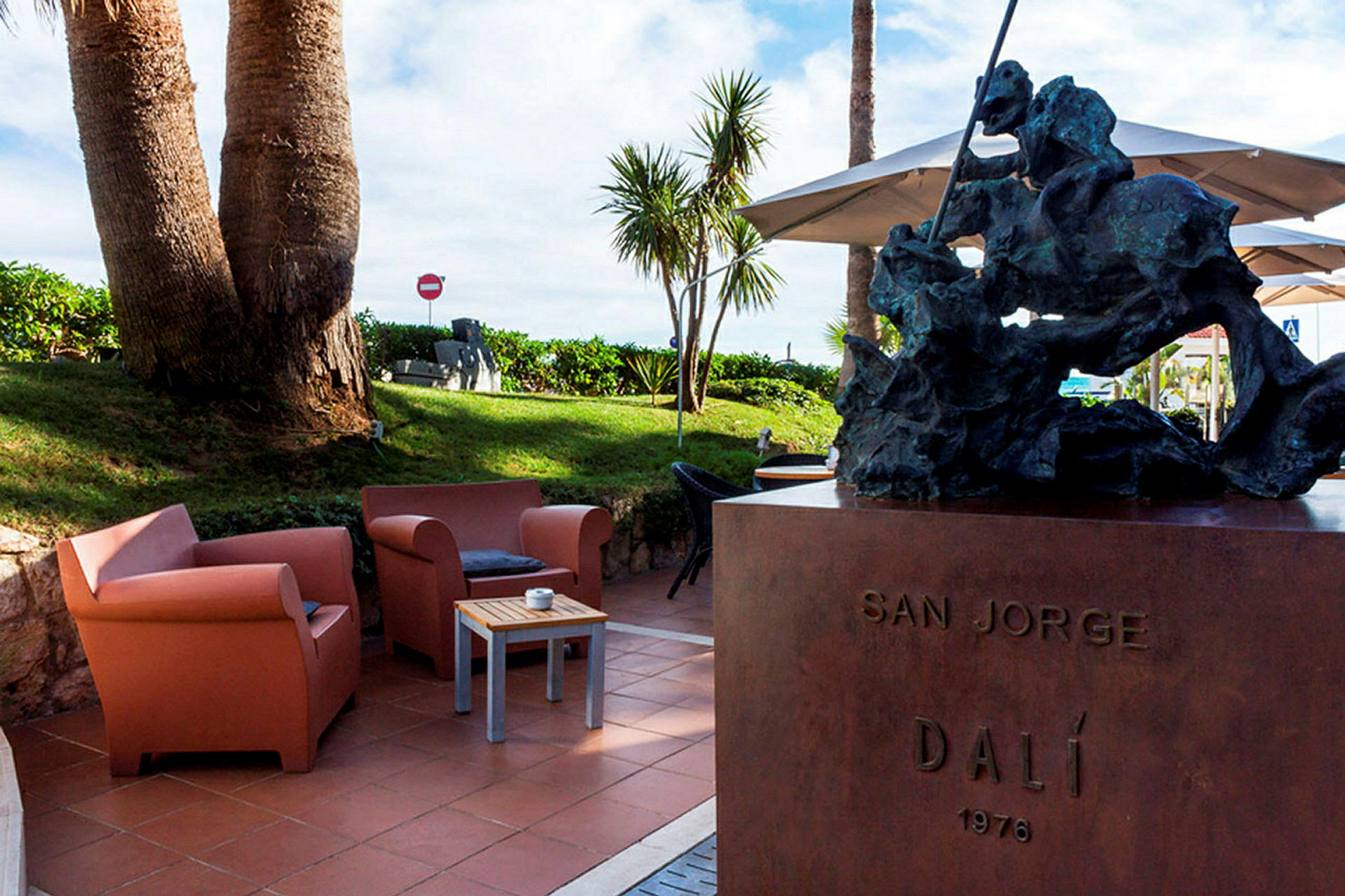 En statue står på en sokkel med teksten 'SAN JORGE DALÍ 1976'. I baggrunden ses en græsplæne med palmer og siddemøbler på en terrasse. (AI-genereret tekst)