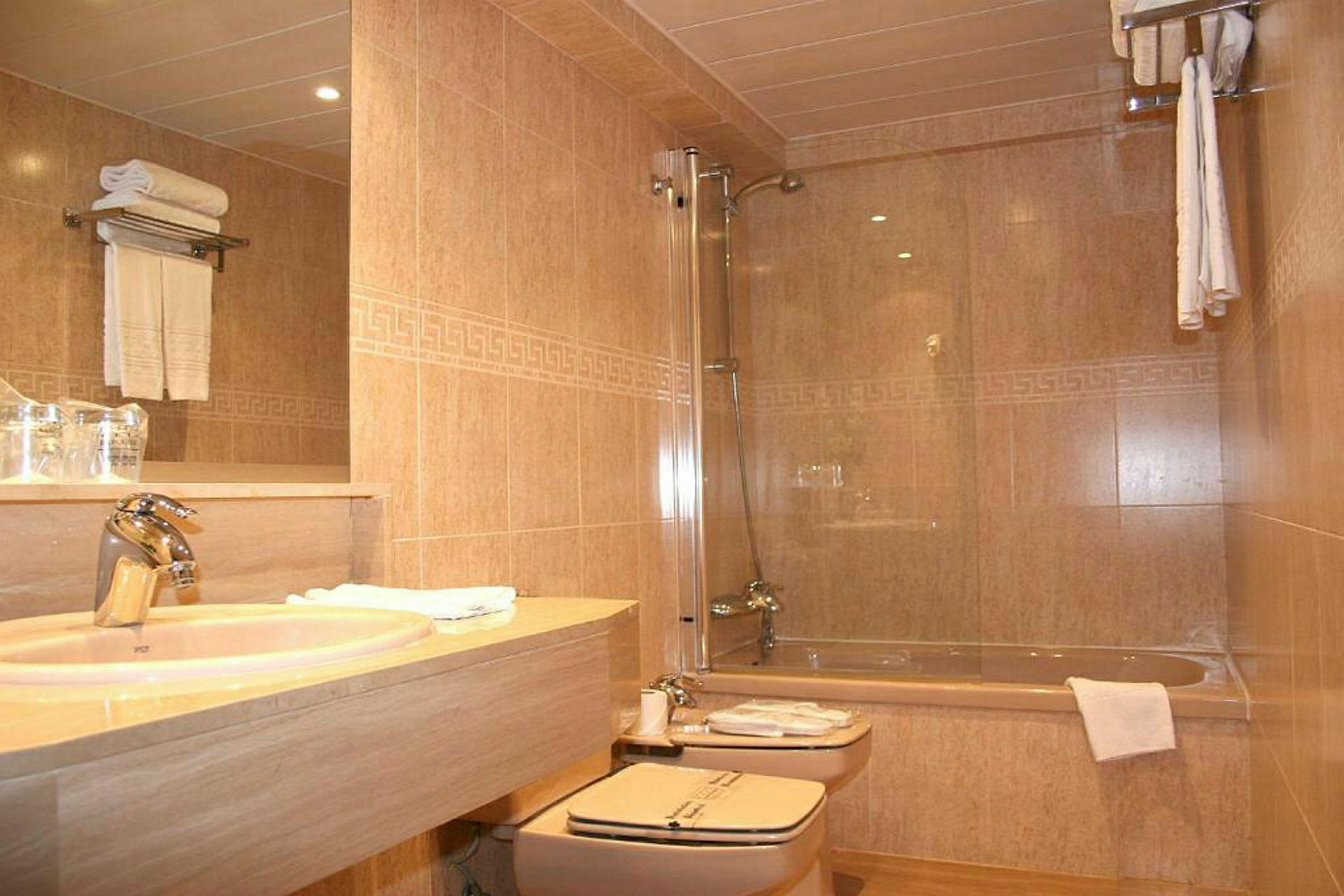 Et badeværelse med håndvask, toilet og badekar. Håndklæder er placeret på en hylde og omkring badekarret. (AI-genereret tekst)