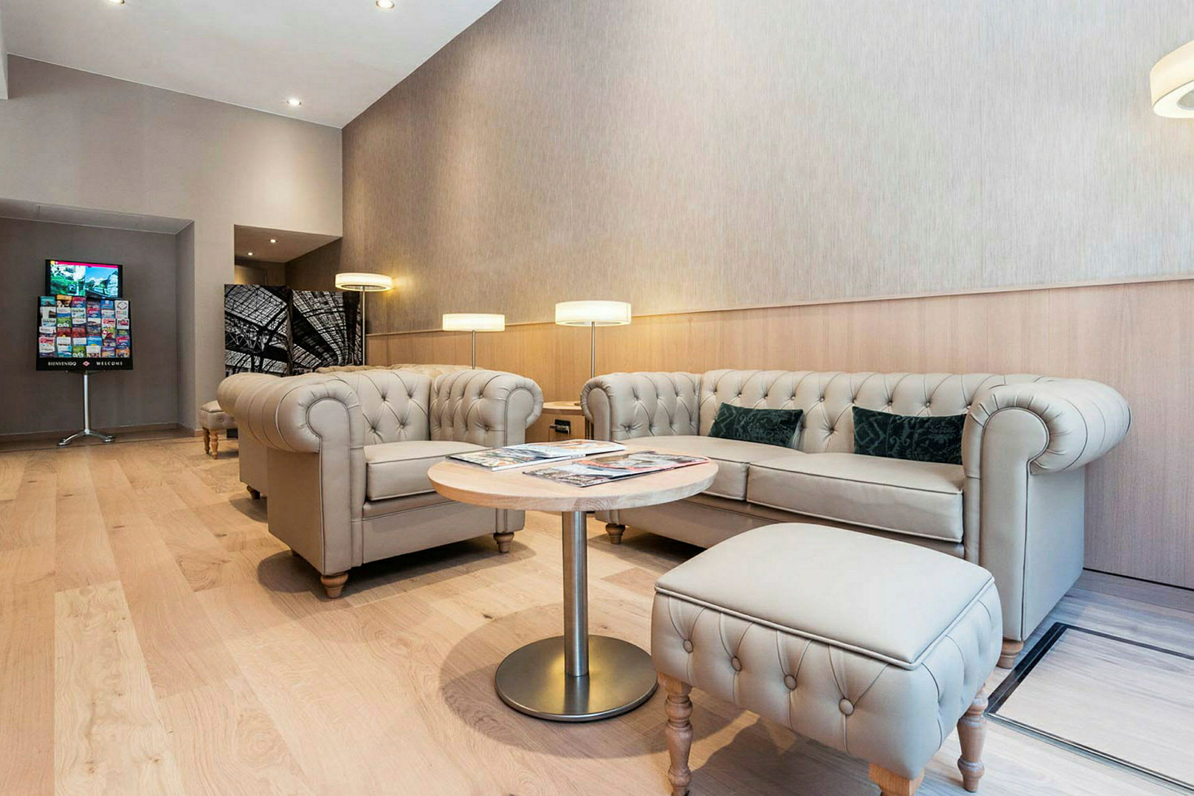 Et moderne loungeområde med beige chesterfieldsofaer og lænestole, et rundt træbord og gulvlamper. Gulvet er af lyst trægulv. (AI-genereret tekst)