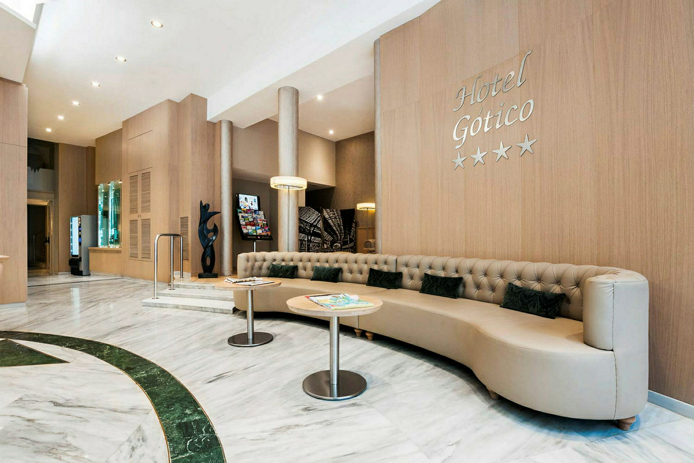 Hotellobby med moderne, lys indretning. En stor, beige sofa med små puder og to runde borde kan ses. Væggene er beklædt med træpaneler, og et skilt med teksten "Hotel Gotico" er synligt. (AI-genereret tekst)