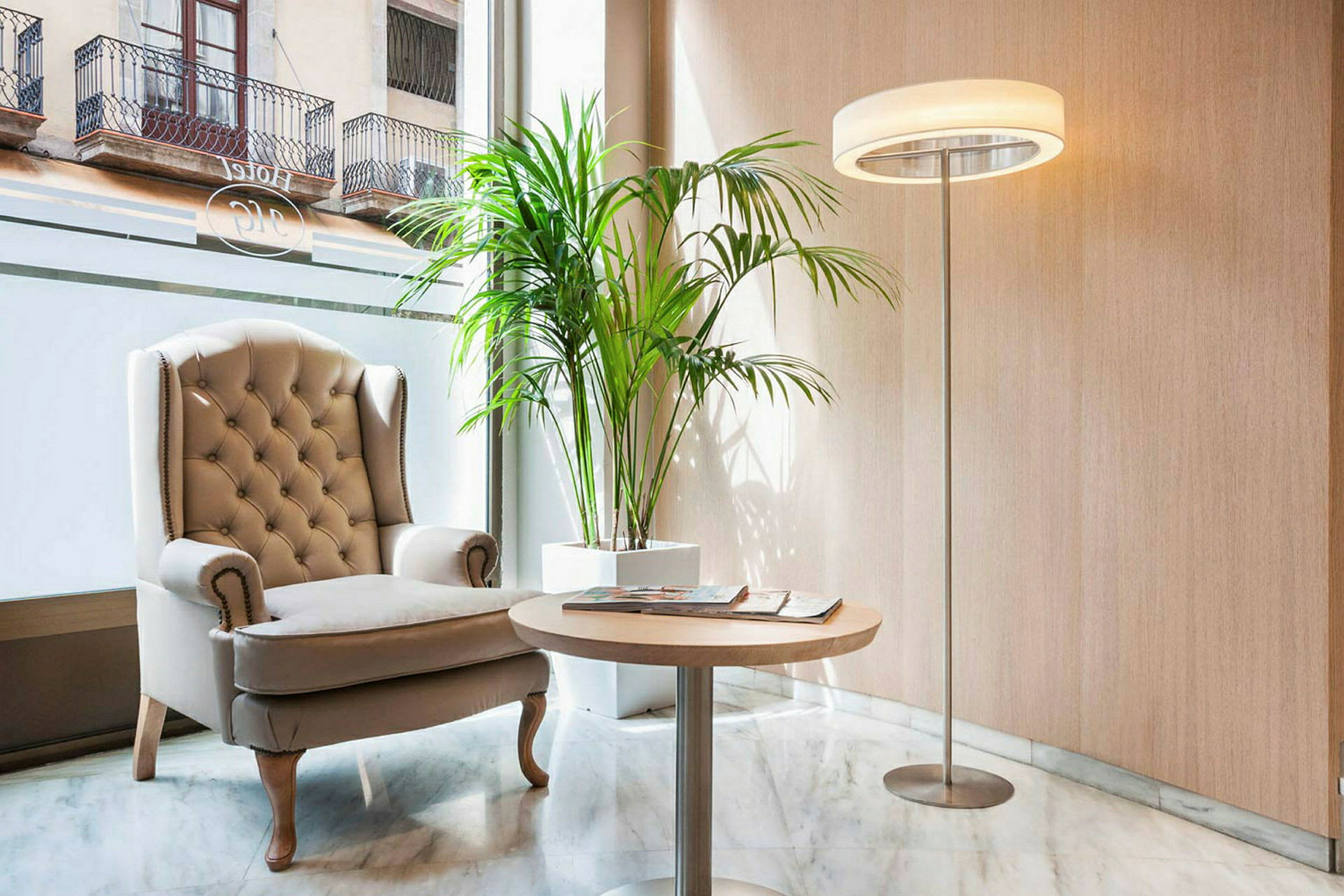 Et rum med en komfortabel beige lænestol, et rundt træbord, en lampe og en grøn plante. Store vinduer i baggrunden. (AI-genereret tekst)