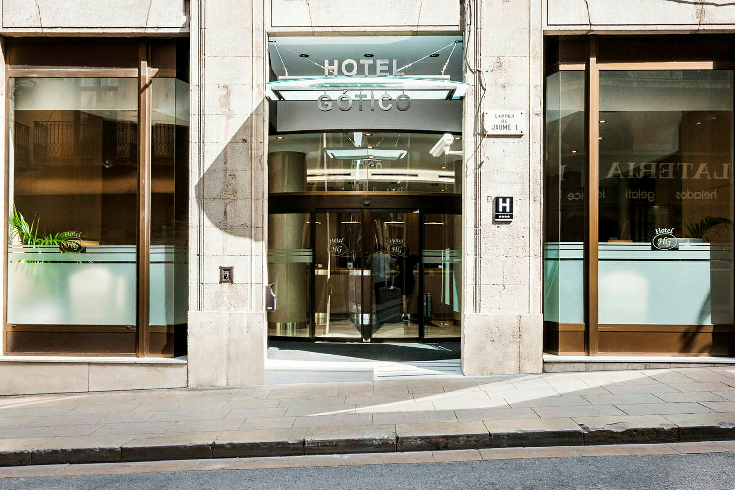 Indgangen til et hotel med glasdøre og hotellets navn synligt ovenover. (AI-genereret tekst)