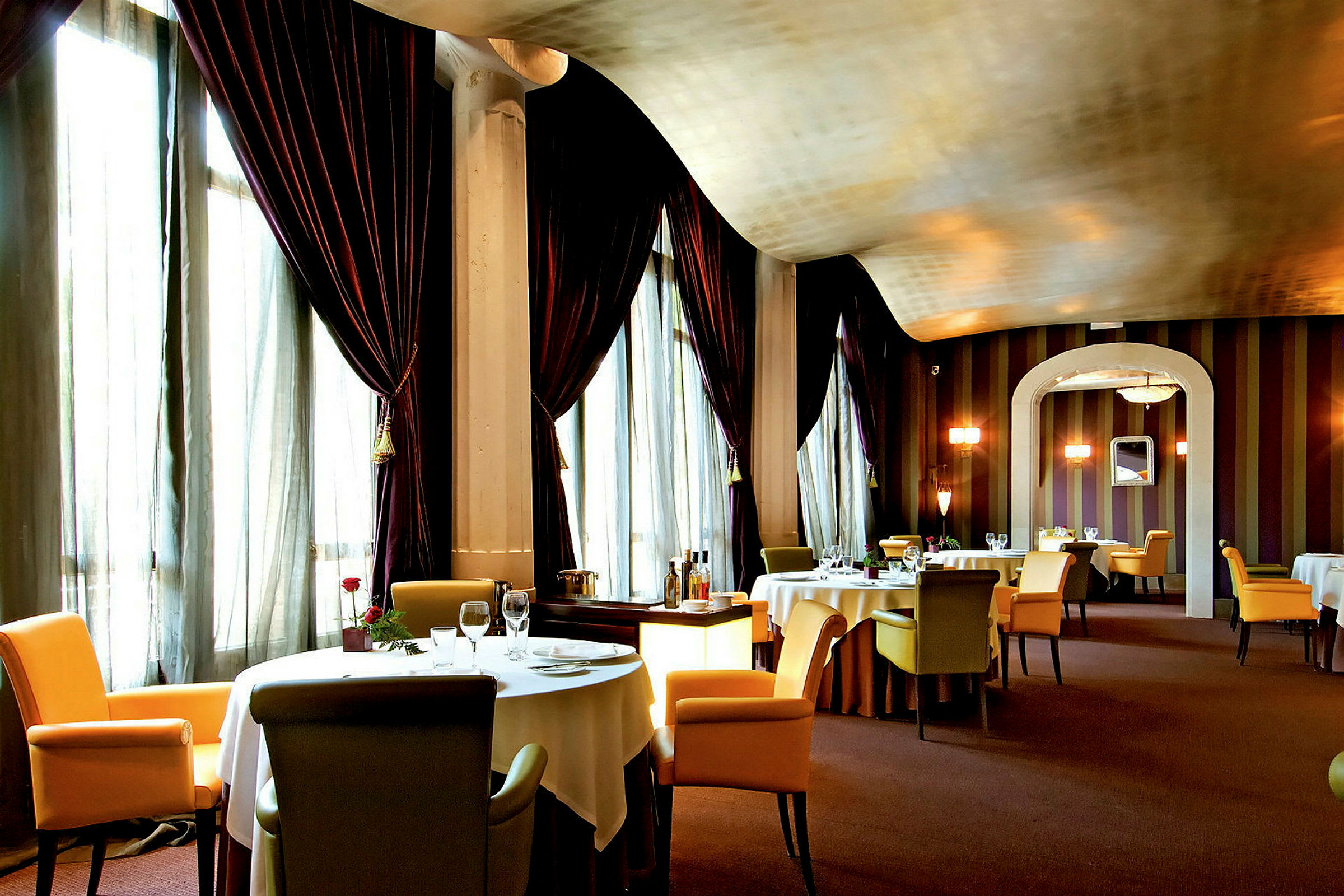 En elegant restaurant med opdækkede borde og komfortable stole, omgivet af store gardiner og dæmpet belysning. (AI-genereret tekst)