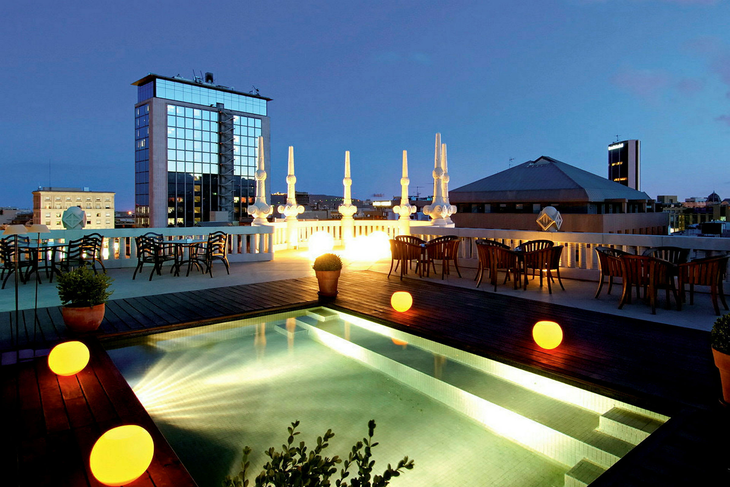 En tagterrasse med en oplyst pool, omgivet af borde og stole samt udsigt over byen i baggrunden under tusmørket. (AI-genereret tekst)